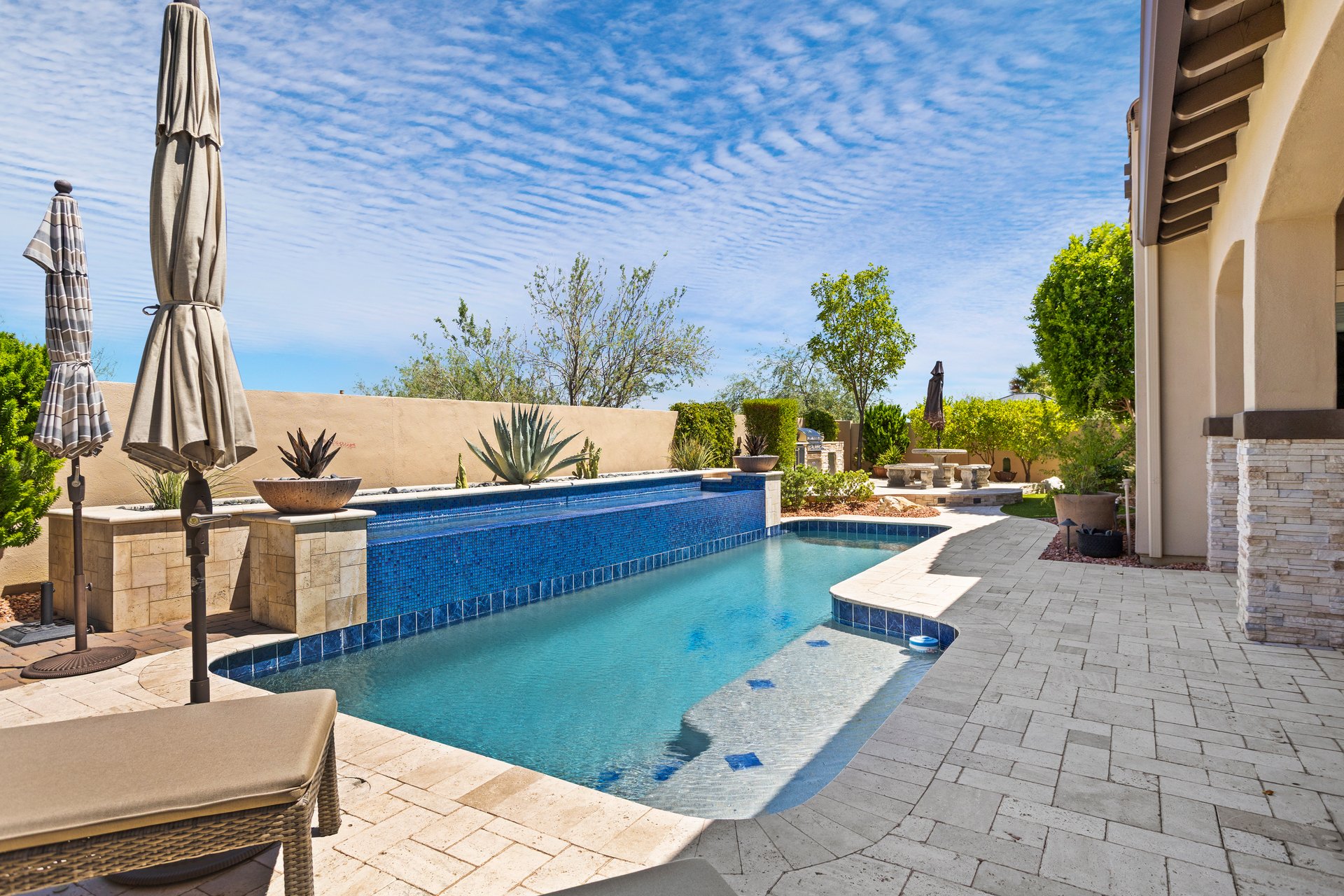 Luxury backyard patio with a swimming pool, spa, stone pavers, umbrellas, and landscaped seating area beside the house.