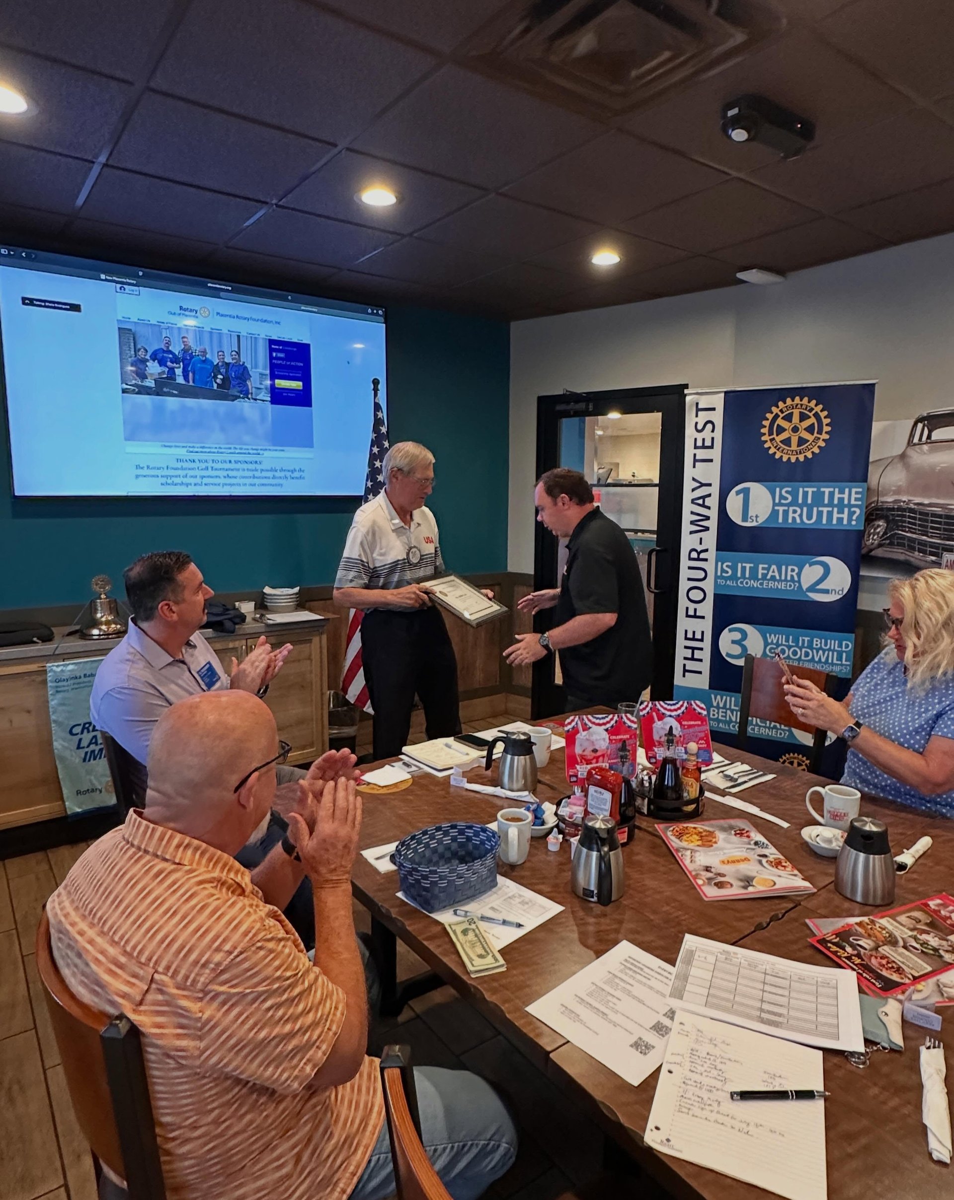 Rotary meeting attendees applaud as a man presents a certificate beside a Four-Way Test banner.