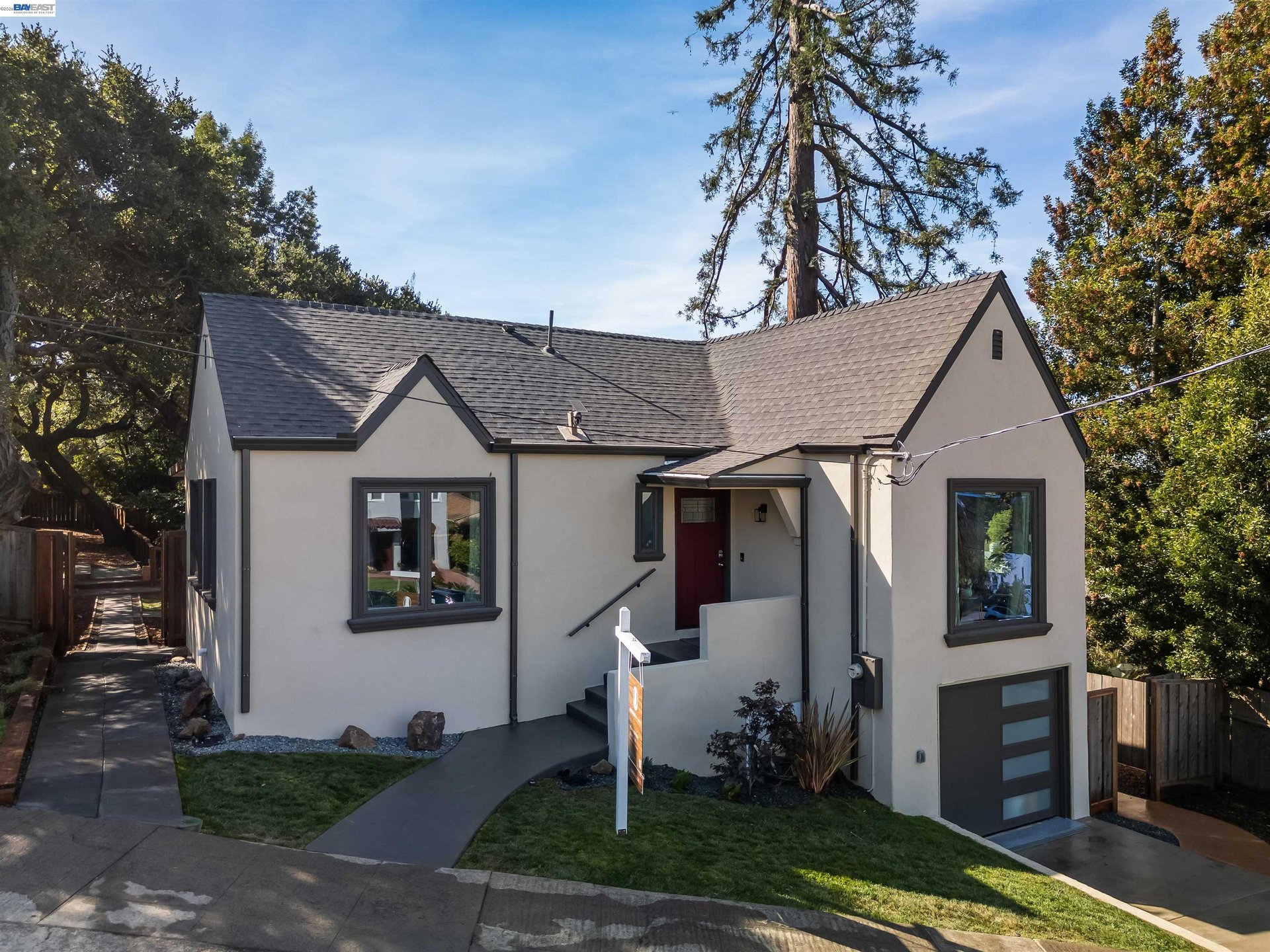 Light-colored house with a gray shingle roof, red entry door, and mature trees.