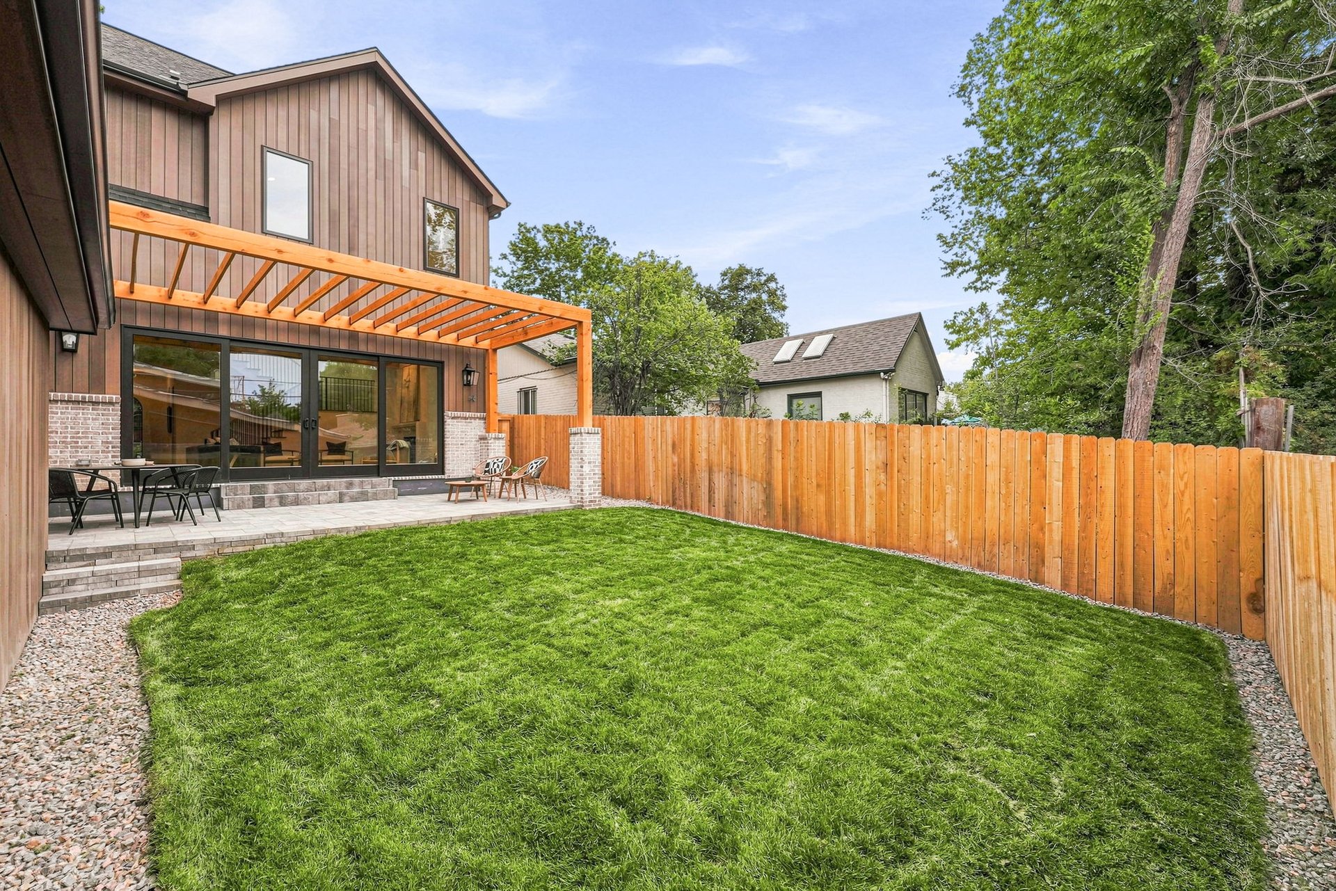Home’s rear exterior with a pergola-covered patio, stone steps, fenced lawn, and mature trees.