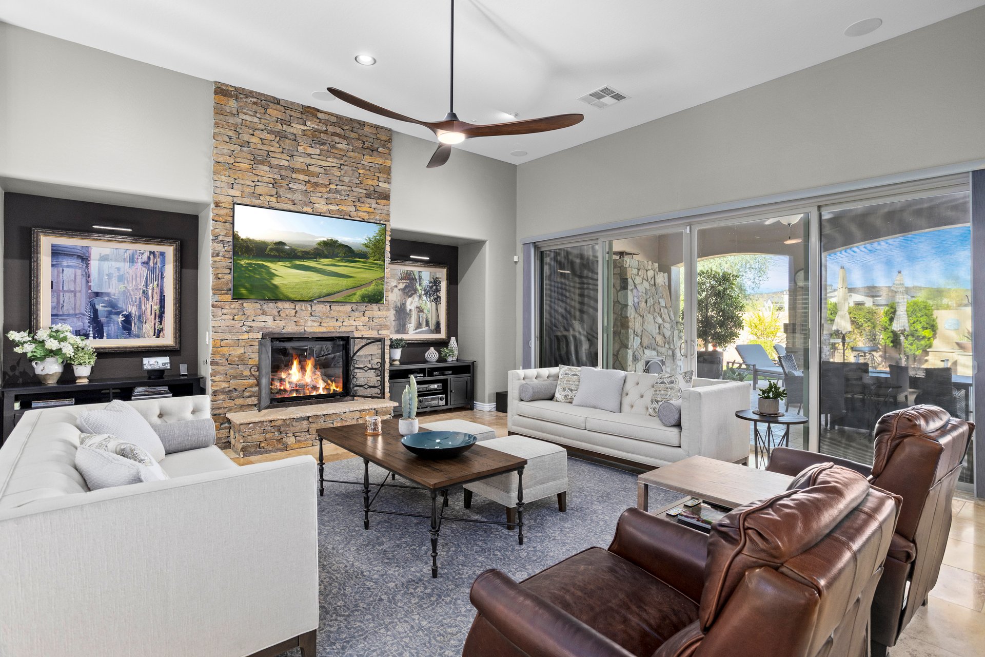 Bright furnished living room with stone fireplace, mounted TV, sofas, armchairs, and sliding glass doors to a patio.