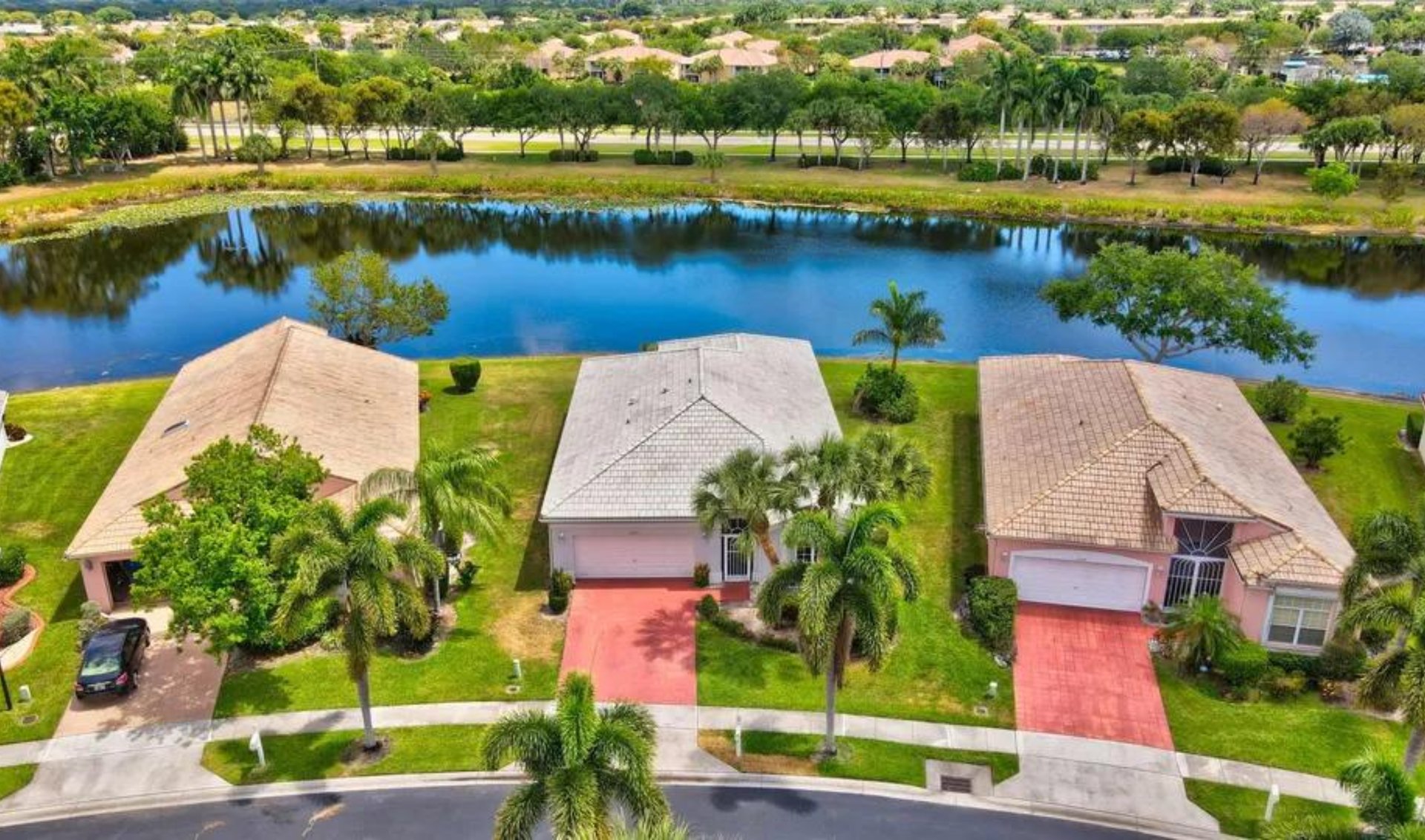 Three homes with red driveways and palm trees beside a community lake.