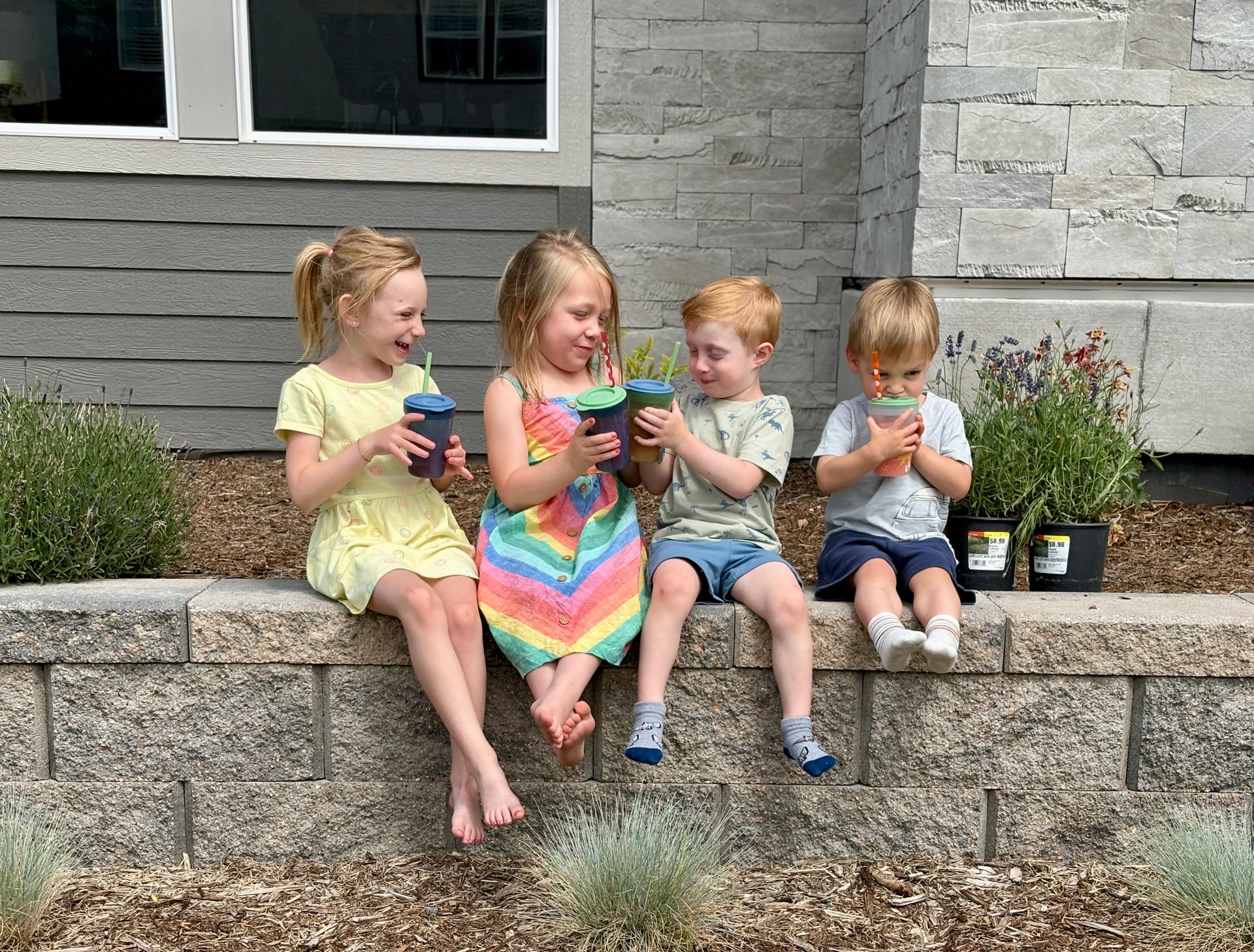 Four children sit outside a home, sharing drinks on a stone garden wall.