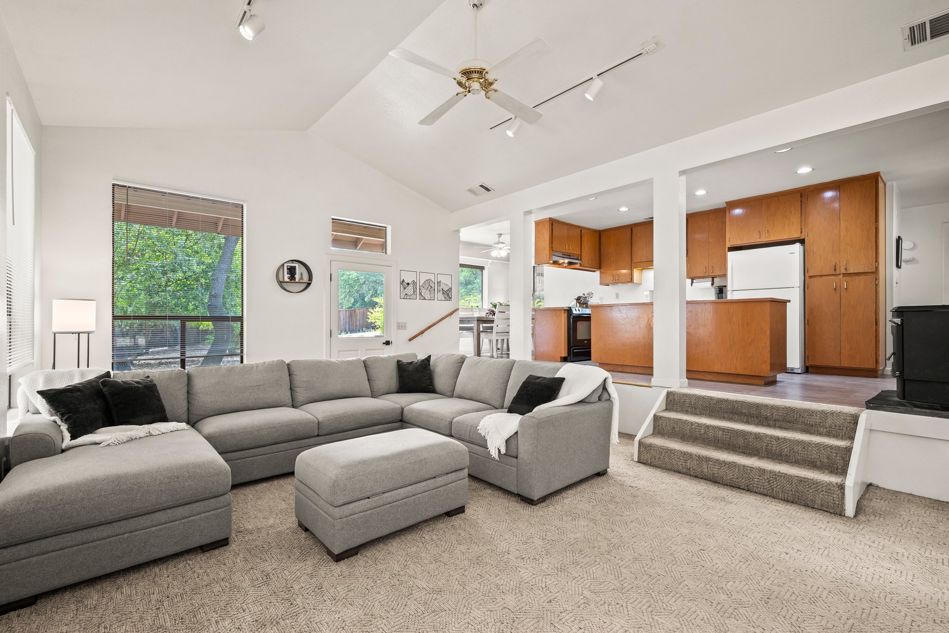 Living room with a gray sectional and ottoman opening onto a kitchen with warm wood cabinets.