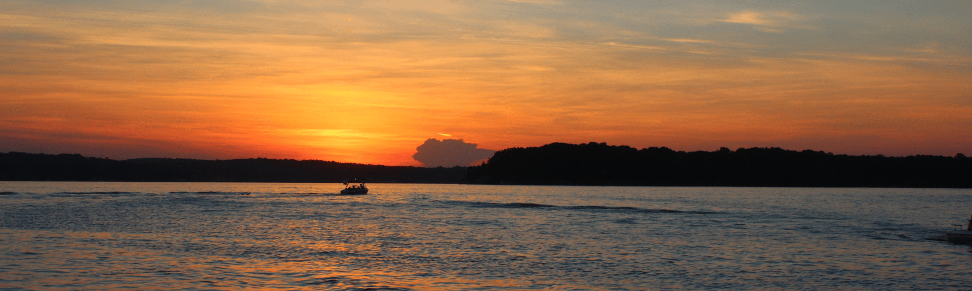 A serene sunset over water, with a boat cruising and silhouettes of trees in the background. Warm hues of orange and pink fill the sky.