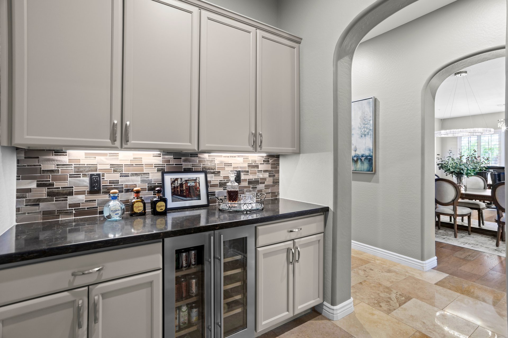 Built-in bar cabinet with upper and lower storage, mosaic tile backsplash, dark countertop, and a small beverage fridge, opening to a dining room through an arched hallway.