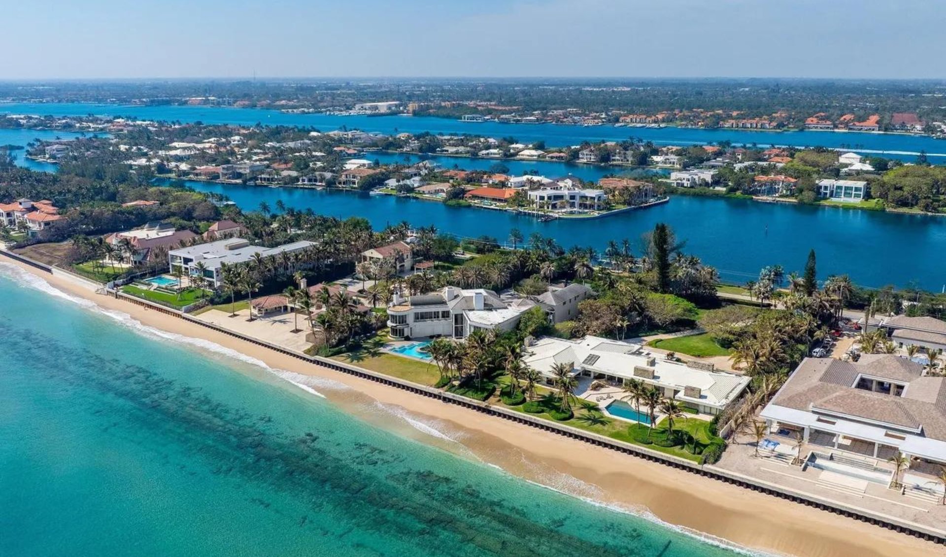 Aerial view of oceanfront homes with private pools, a sandy beach, and a wide waterway beyond.