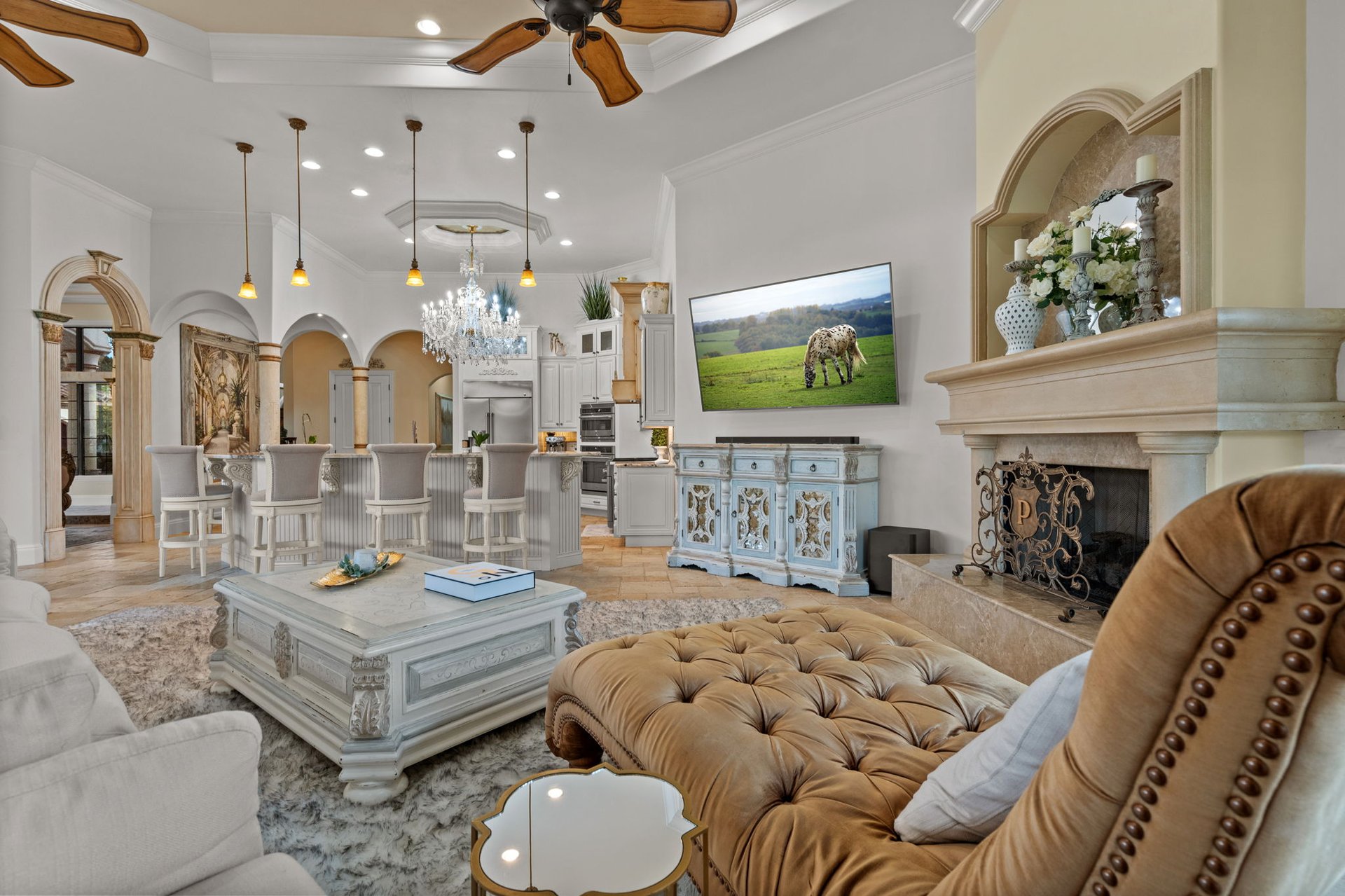 Living room with a stone fireplace, tufted tan ottoman, and adjoining kitchen.