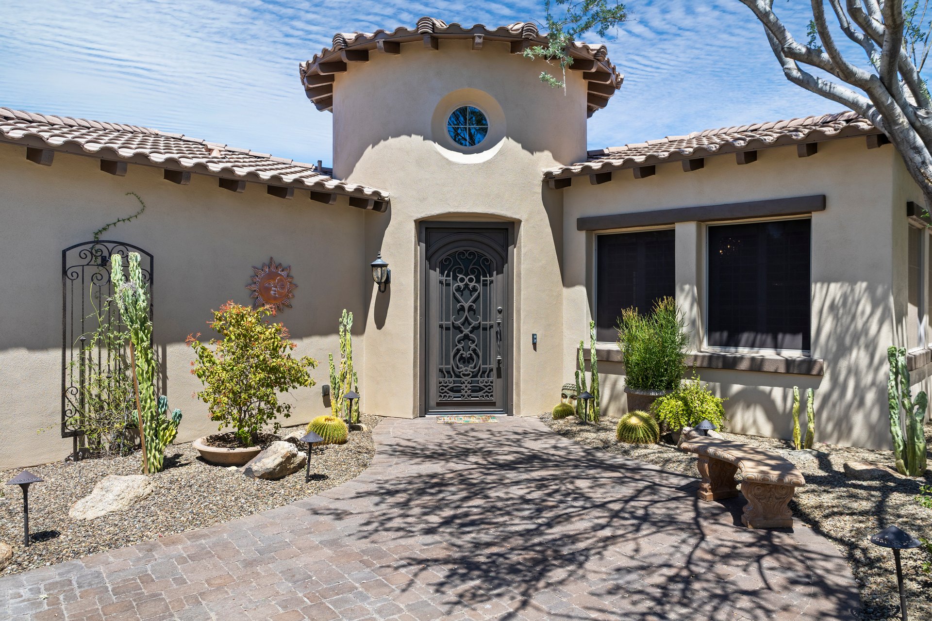 Front entrance of a stucco house with a tiled roof, arched tower detail, iron security door, desert landscaping, and brick paver walkway.