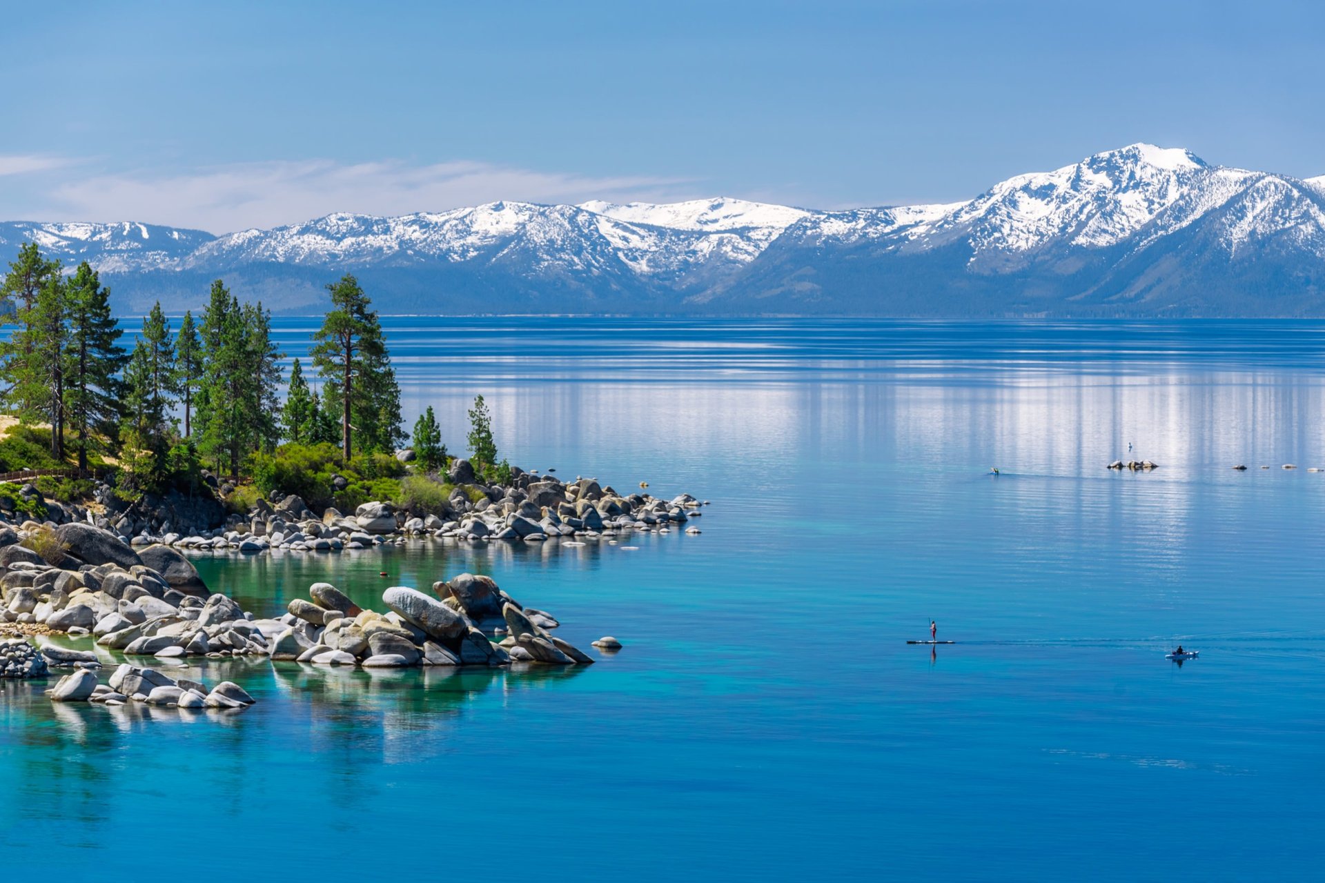 Lake Tahoe with a rocky pine shoreline, snow-covered mountains, and paddleboarders on the water.