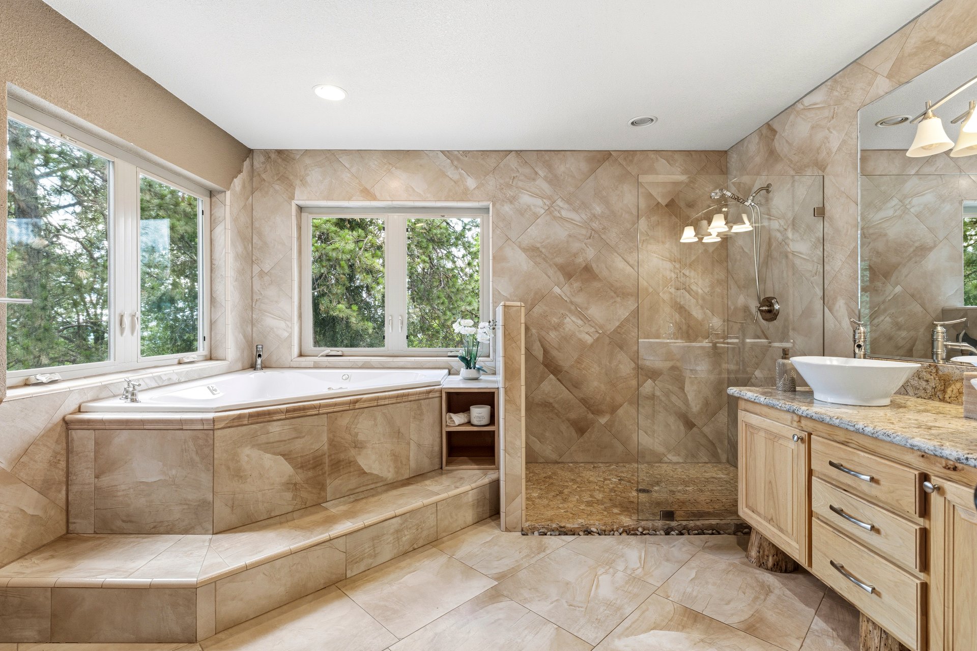 Tiled bathroom with a raised corner soaking tub, glass shower, and wood vanity beneath large windows.