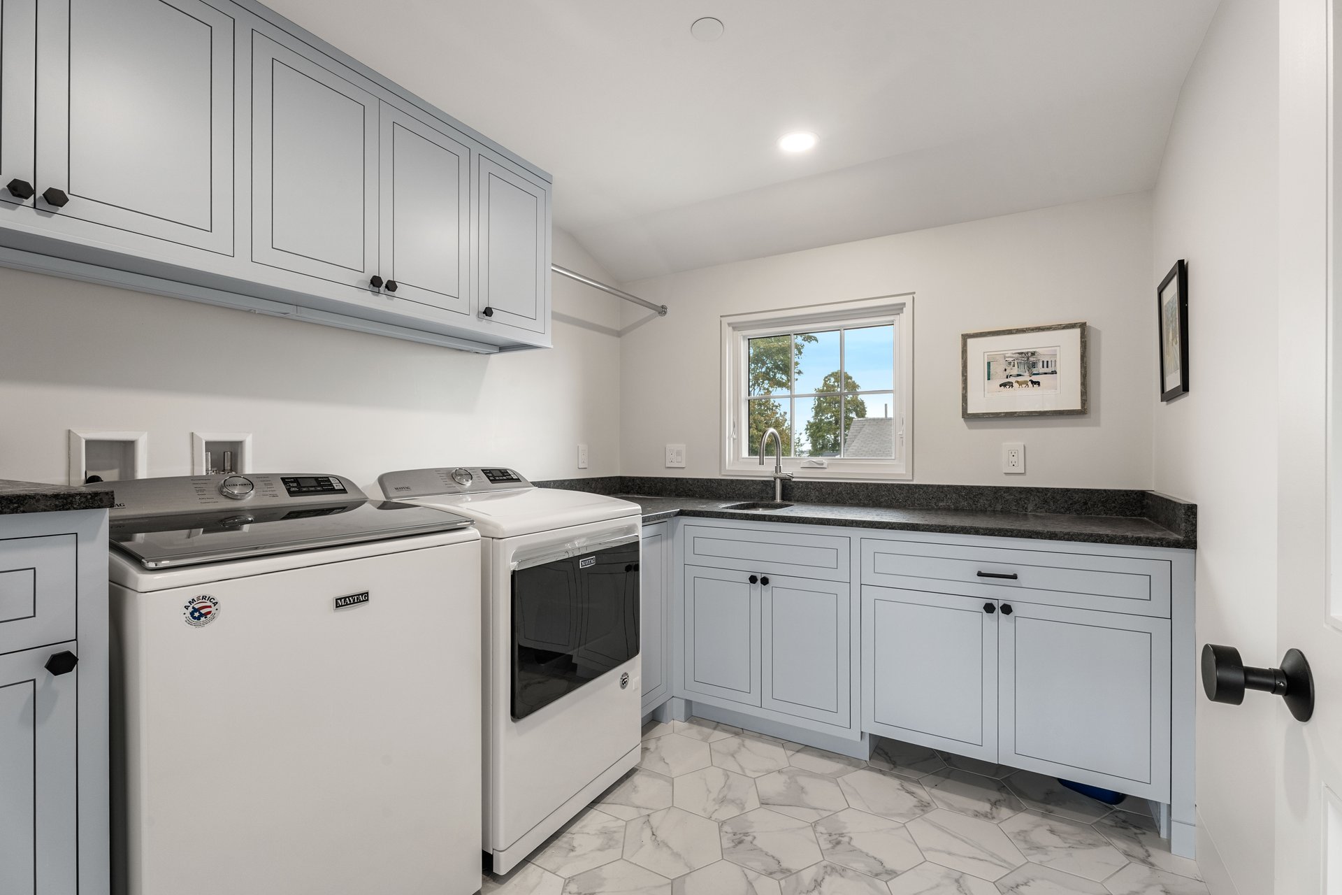 Laundry room with washer and dryer, pale blue-gray cabinetry, dark countertops, and marble-look tile floor.
