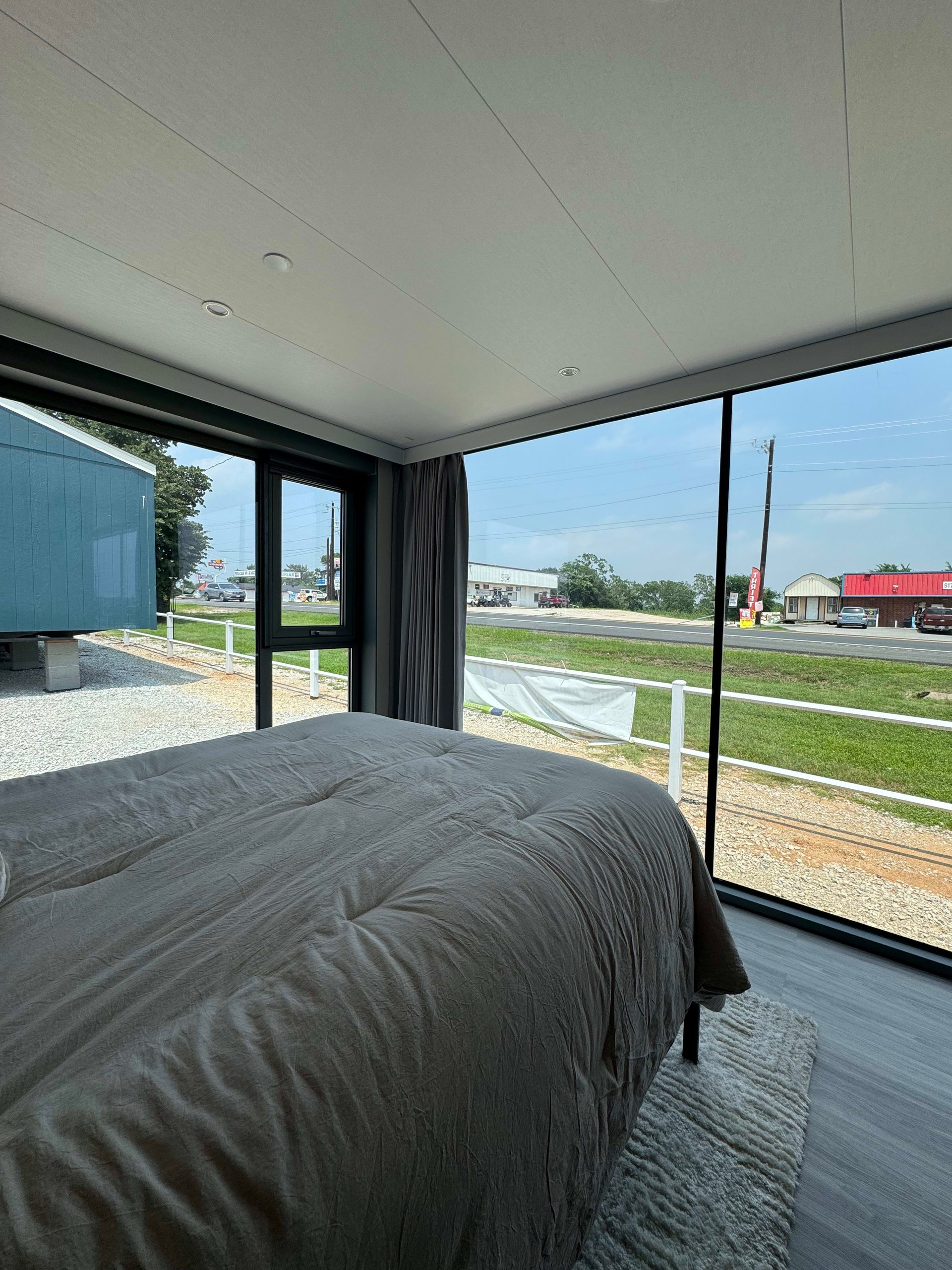 Tiny-home bedroom with a gray-covered bed and floor-to-ceiling windows overlooking the roadside.
