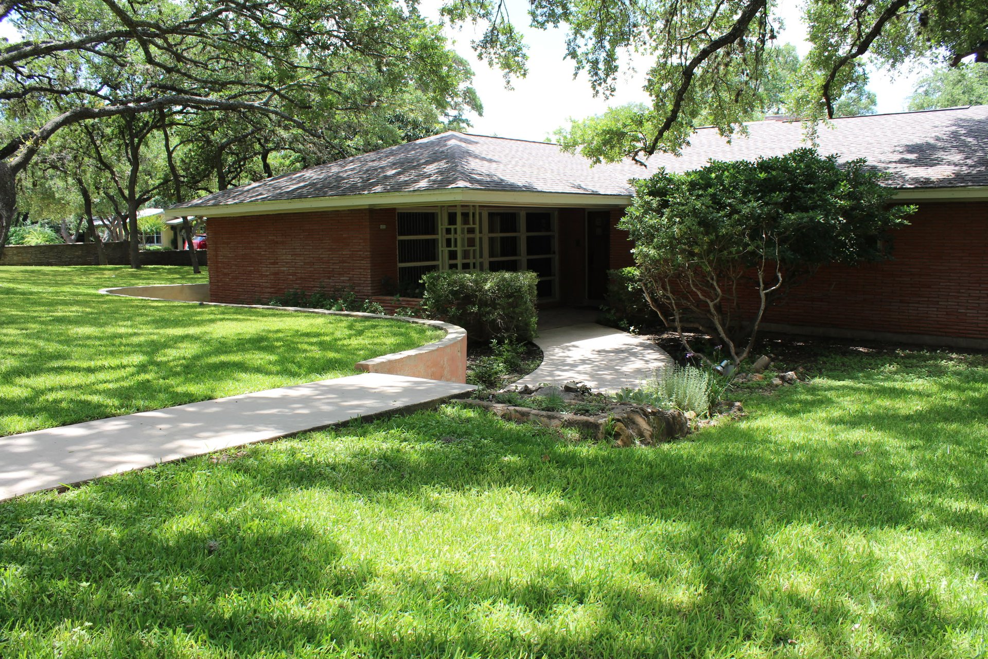 Single-story brick home behind a curved walkway, shaded by mature trees.