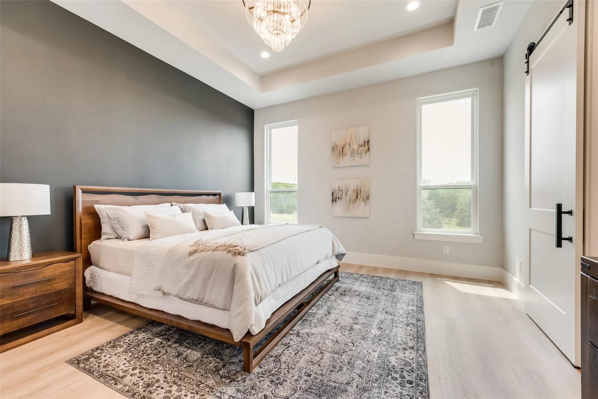 Staged bedroom with a wood bed, charcoal accent wall, chandelier, and two tall windows.