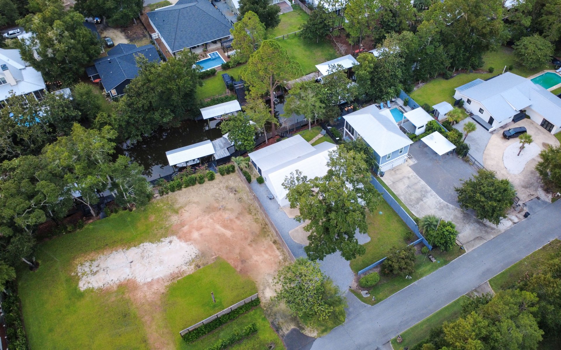 Aerial view of homes along a tree-lined canal, with pools and an undeveloped lot nearby.