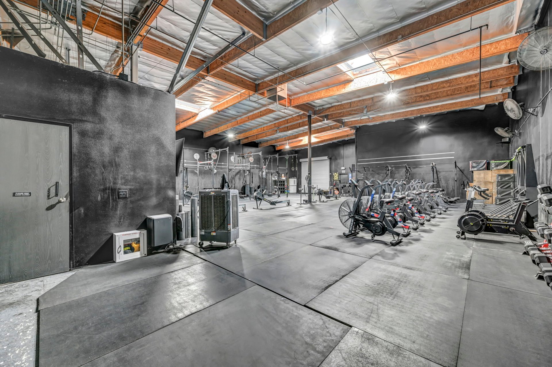 Commercial gym with weight racks, rows of cardio machines, and exposed timber ceiling beams.
