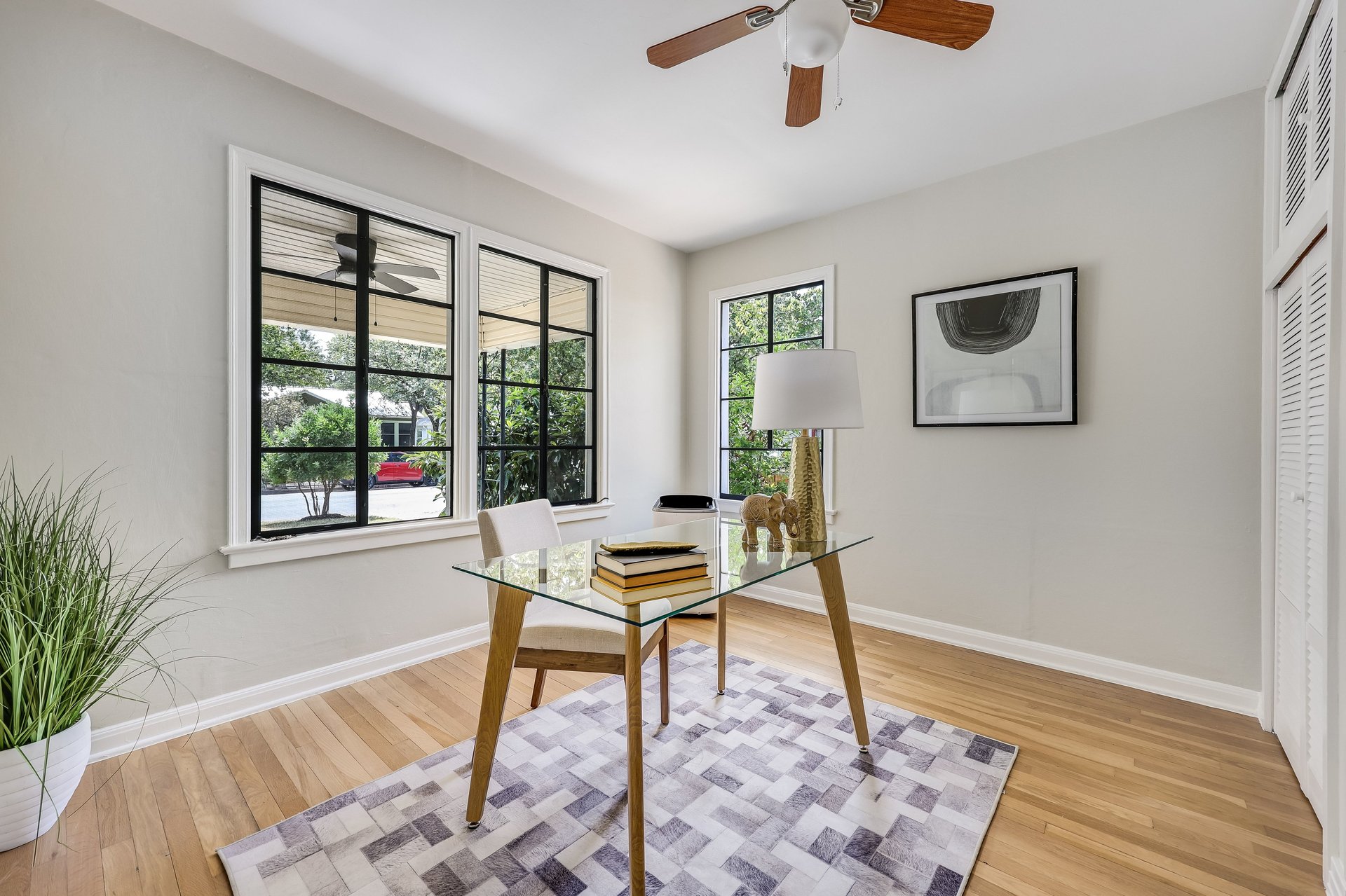 Staged home office with a glass desk, geometric rug, and large black-framed windows.