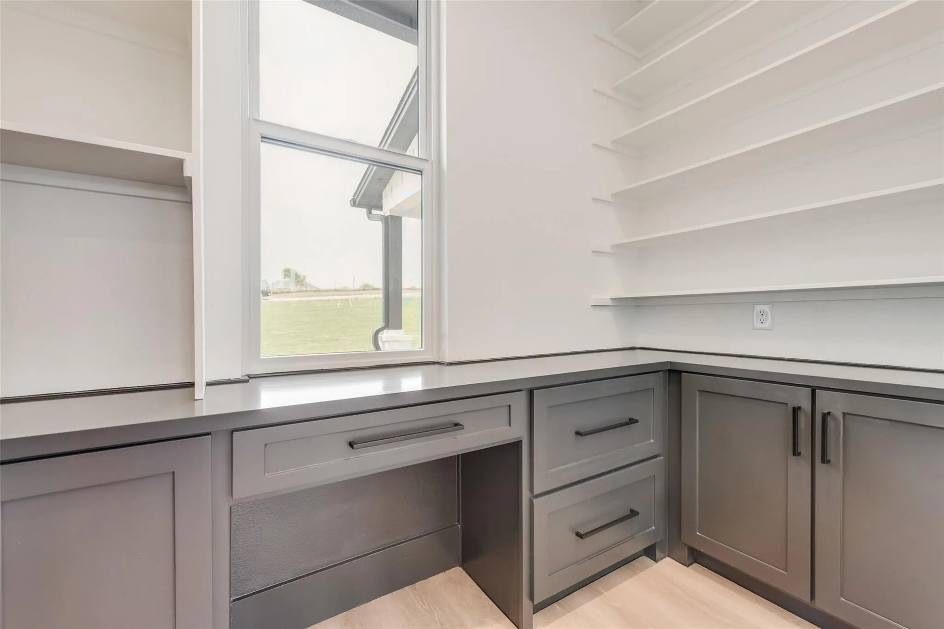 Built-in counter with gray cabinets and open shelves beside a window overlooking a lawn.