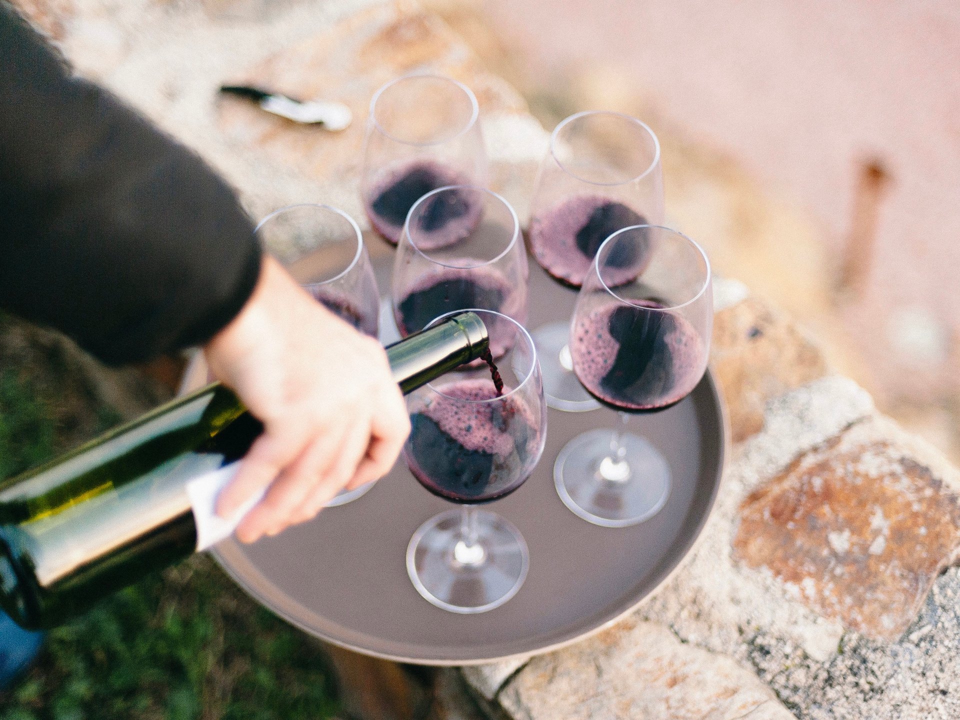 Red wine is poured into a glass surrounded by other wine glasses on a tray.
