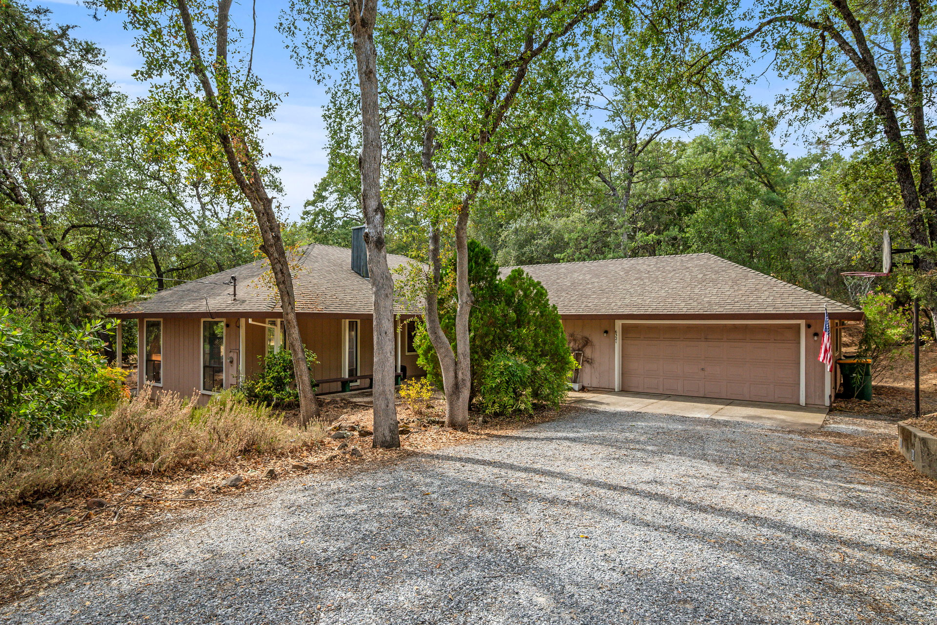 Single-story house with a two-car garage, mature trees, and a broad gravel driveway.