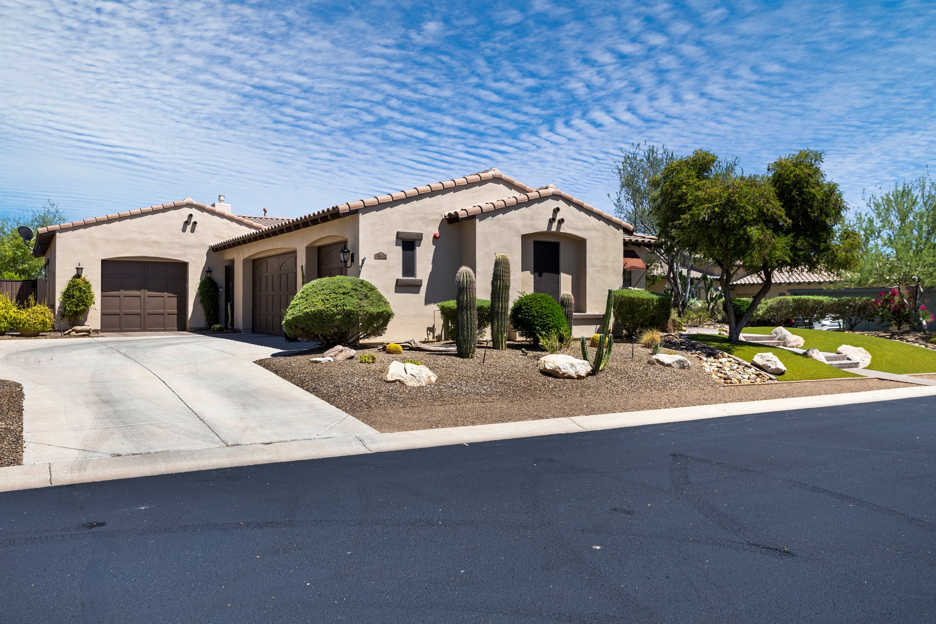 Front exterior of a beige stucco house with tiled roof, driveway, garage, desert landscaping, and a bright blue sky.