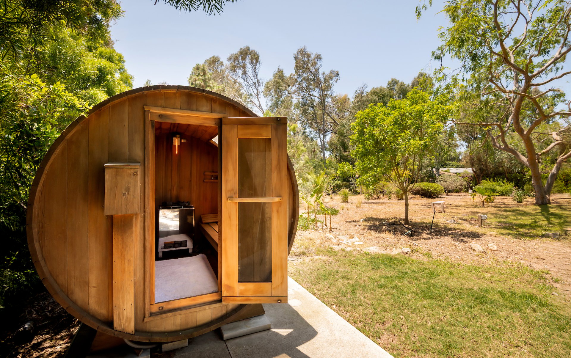 Wooden barrel sauna with its door open in a sunny, tree-lined yard.