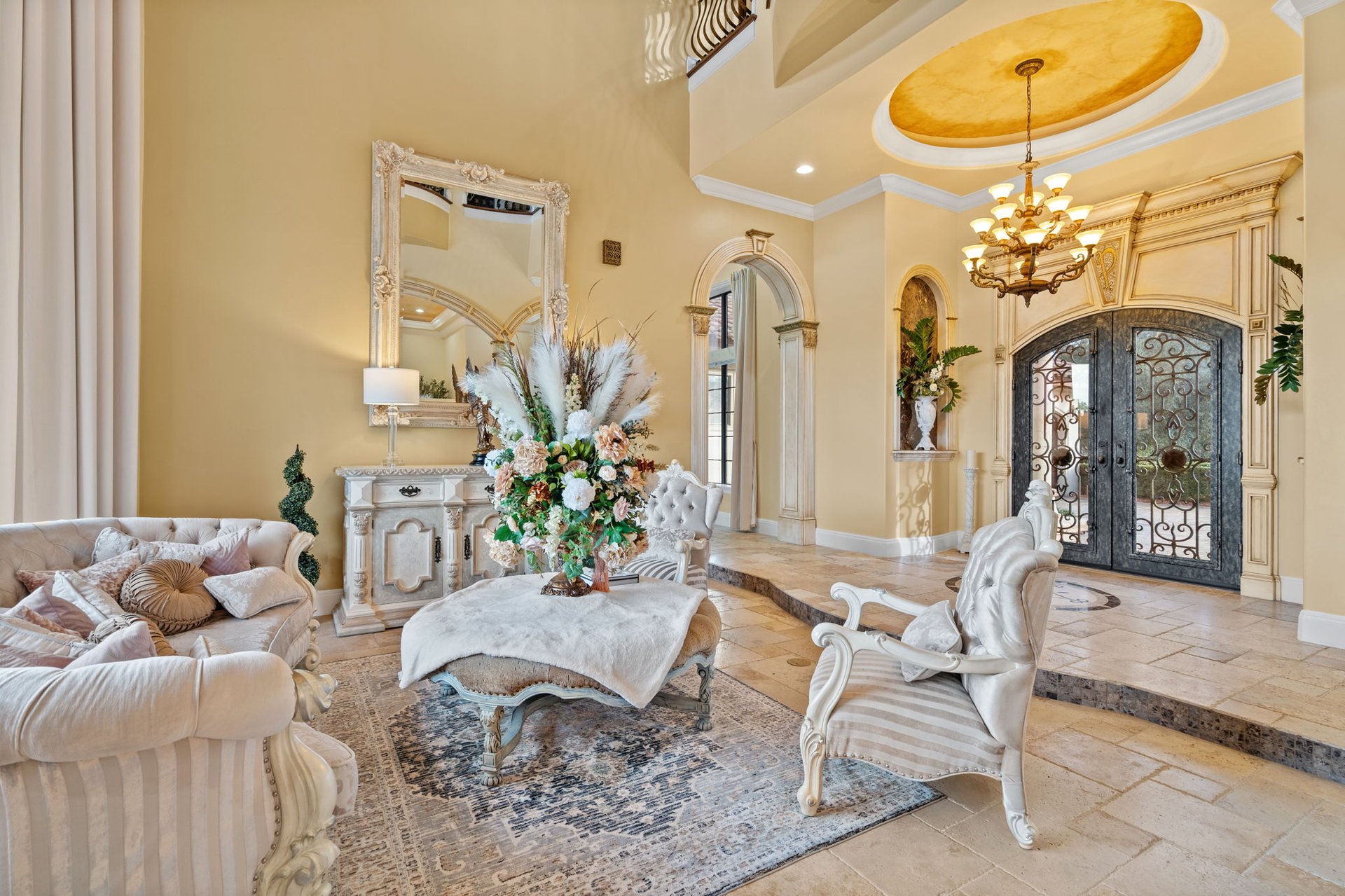 Formal living room with cream seating, carved fireplace mantel, wrought-iron entry doors, and ornate chandelier.