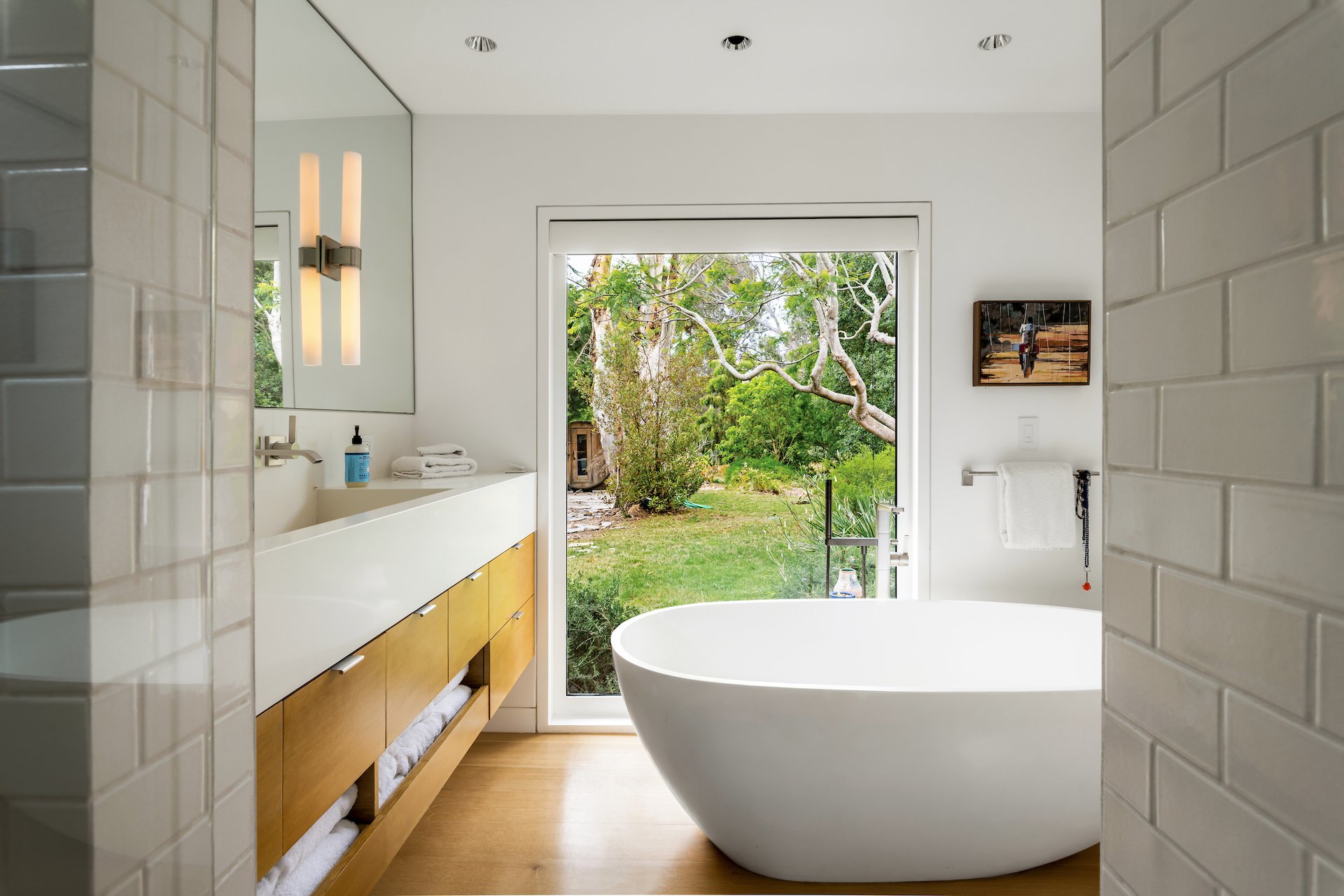 Contemporary bathroom with a freestanding tub, wood vanity, and glass door opening to a leafy garden.