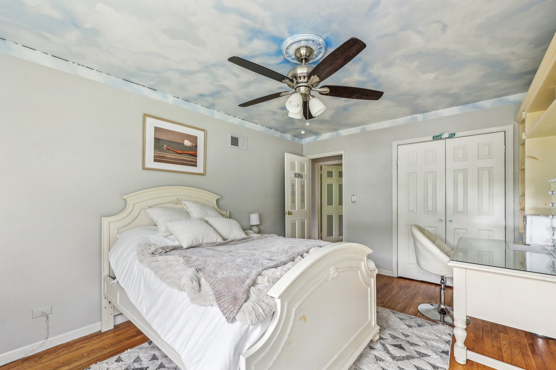 Furnished bedroom with an ornate white bed, cloud-painted ceiling, and dark hardwood floors.