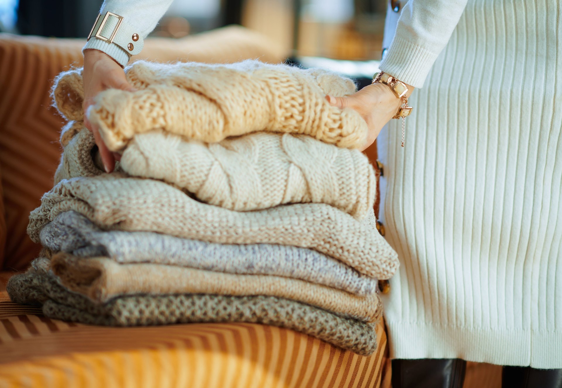 Person arranging folded knit sweaters in warm neutral colors on a striped chair.
