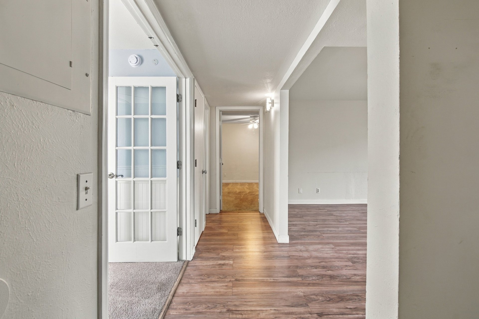 Hallway with wood-look flooring, a glass-paneled door, and an adjoining empty living area.