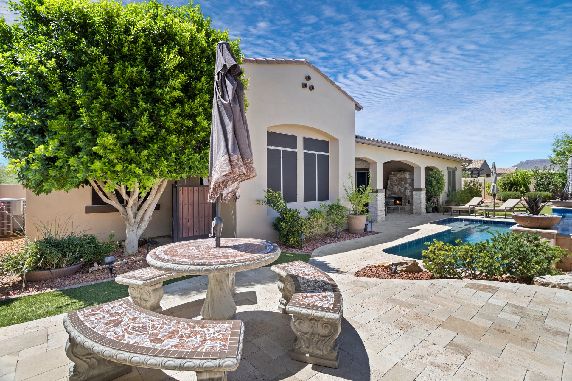 A landscaped backyard patio with a pool, outdoor dining table, lounge chairs, and a covered fireplace area beside a stucco home.