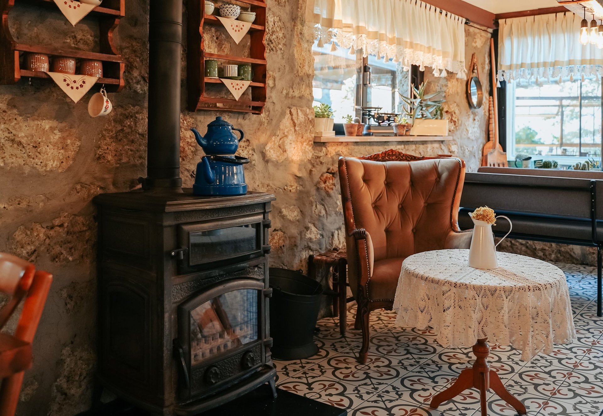 Rustic stone-walled interior with a wood stove, blue kettle, leather armchair, and patterned tile floor.