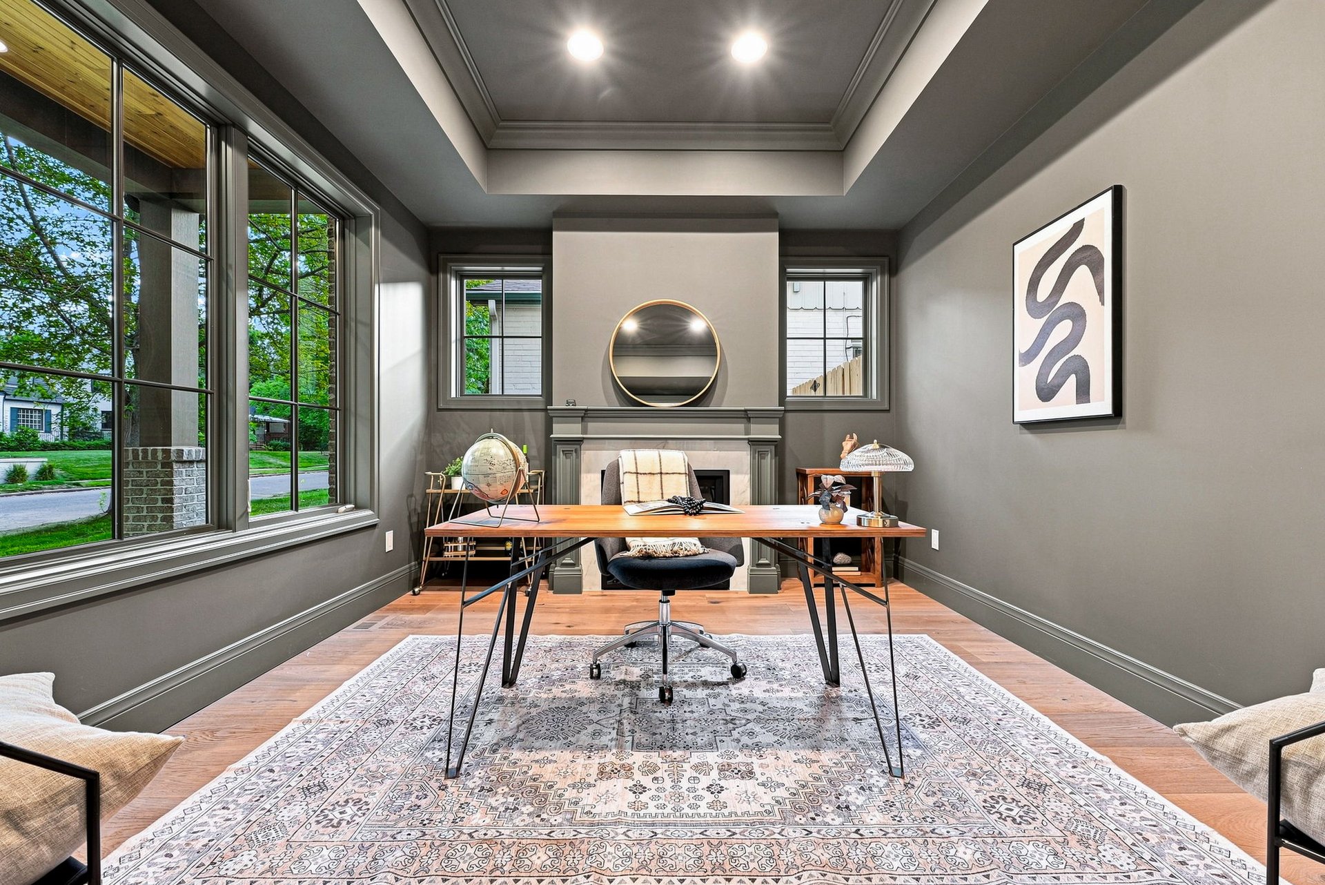 Furnished home office with a wooden desk, fireplace, round mirror, and large windows.