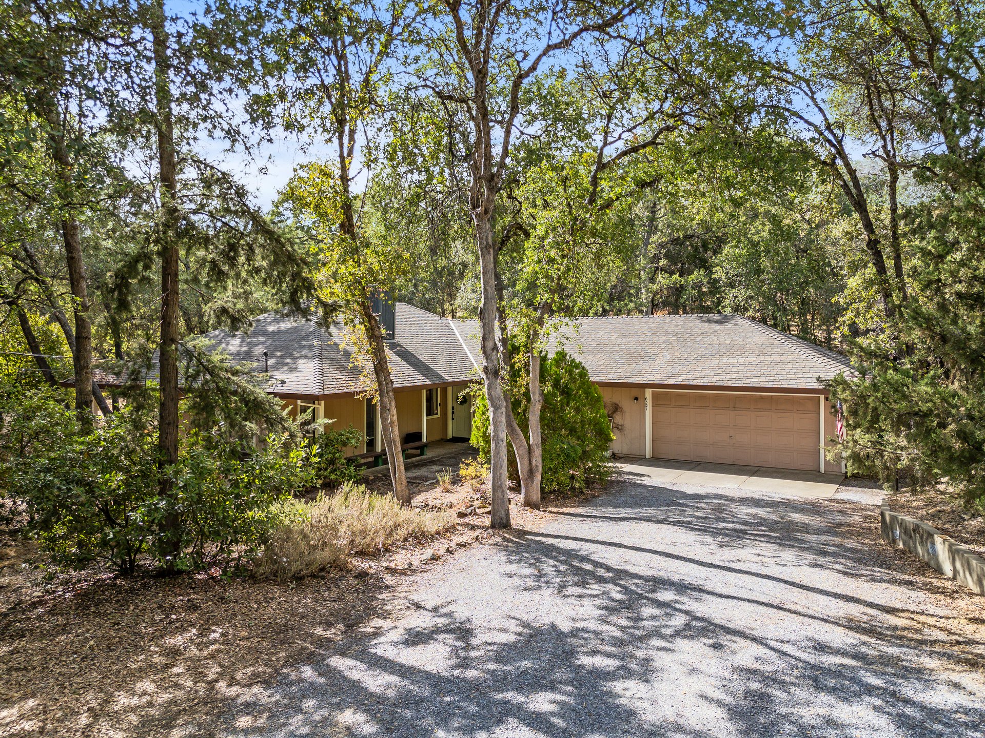 Single-story home with a two-car garage and gravel driveway beneath mature trees.