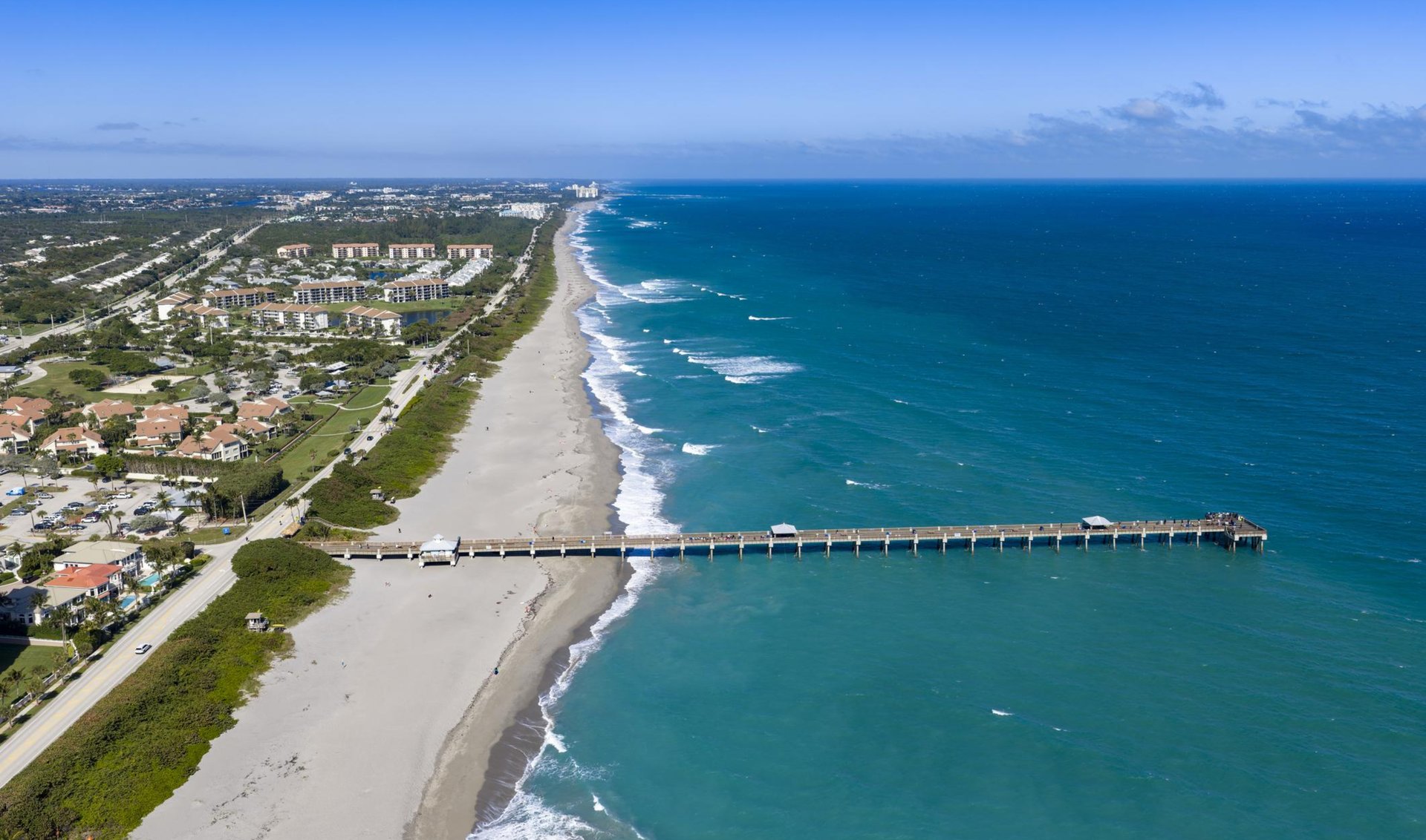 Aerial coastline with a sandy beach, oceanfront homes, and a long pier.