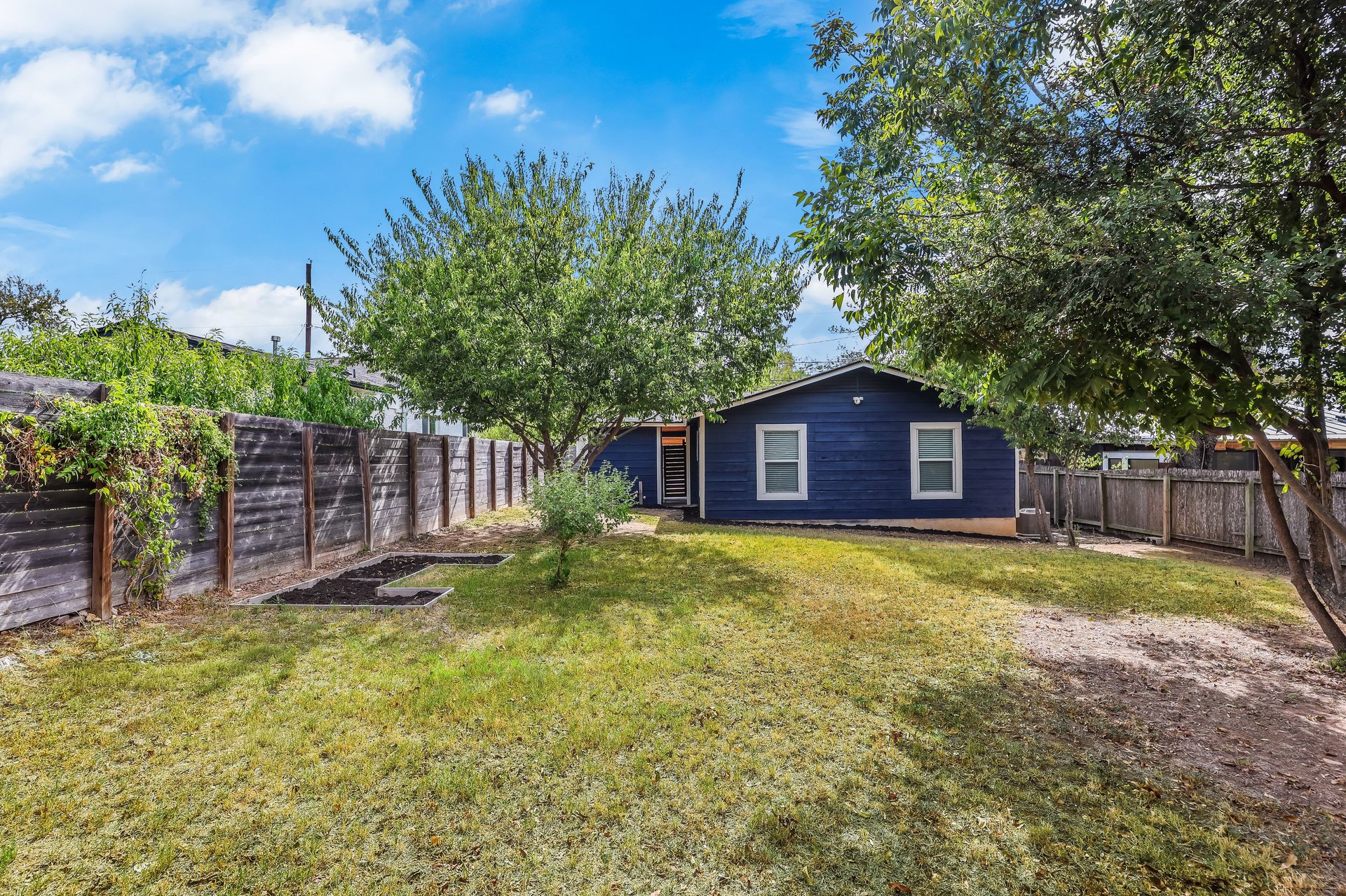 Blue single-story home behind a grassy backyard with mature trees and wooden privacy fencing.