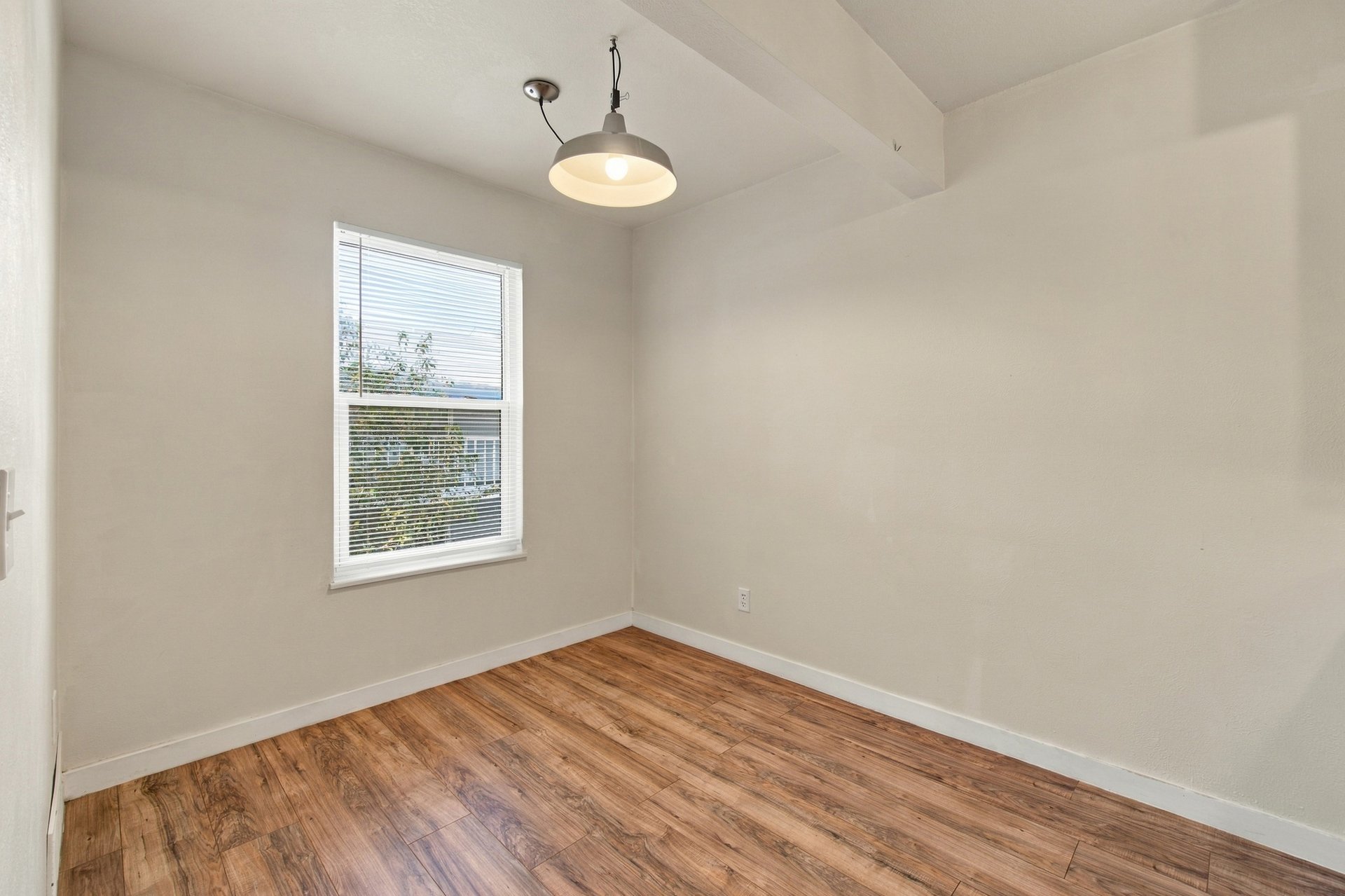 Empty room with wood-look flooring, a window with blinds, and a hanging ceiling light.