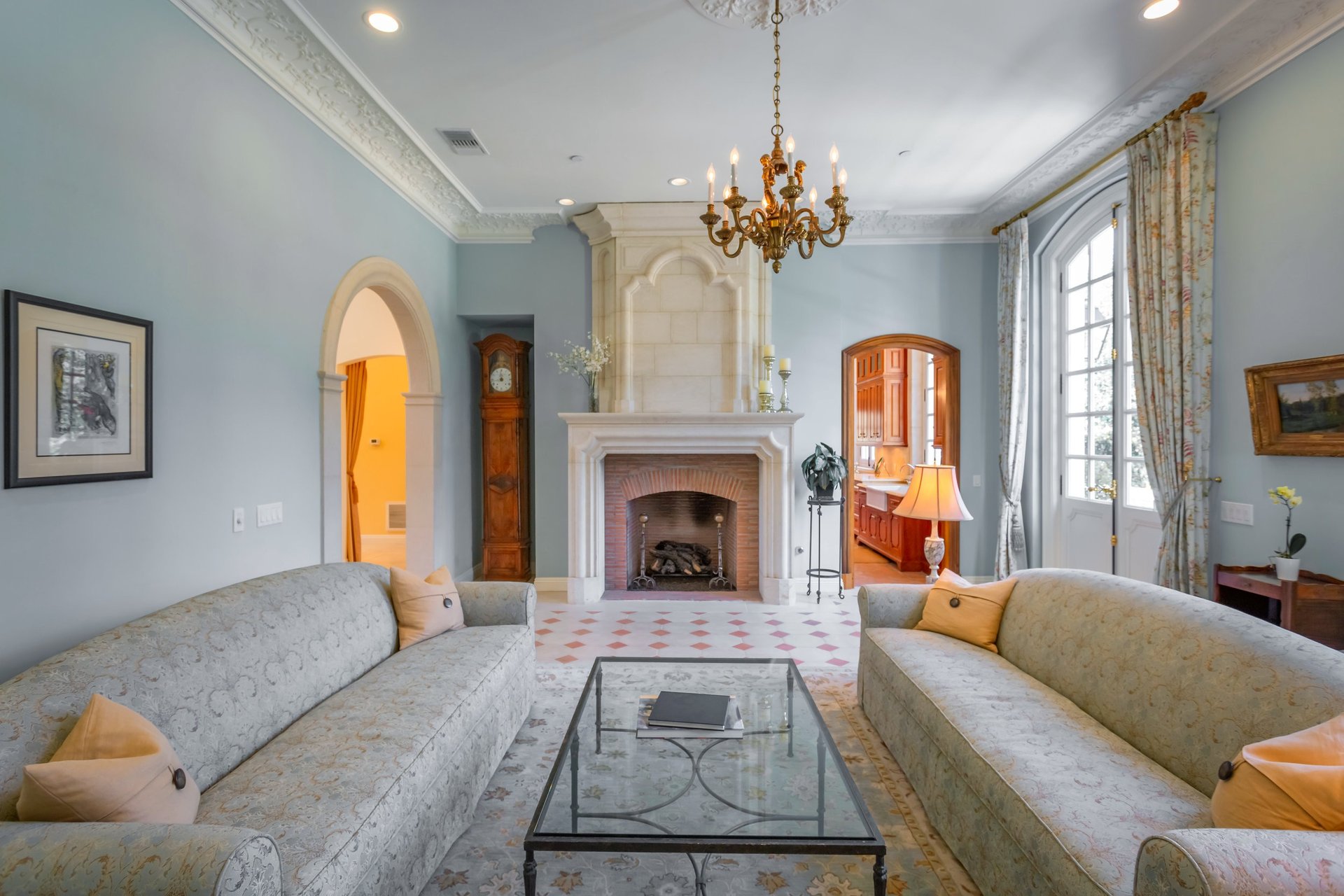 Living room with a carved stone fireplace, ornate chandelier, arched doorways, and tall French doors.