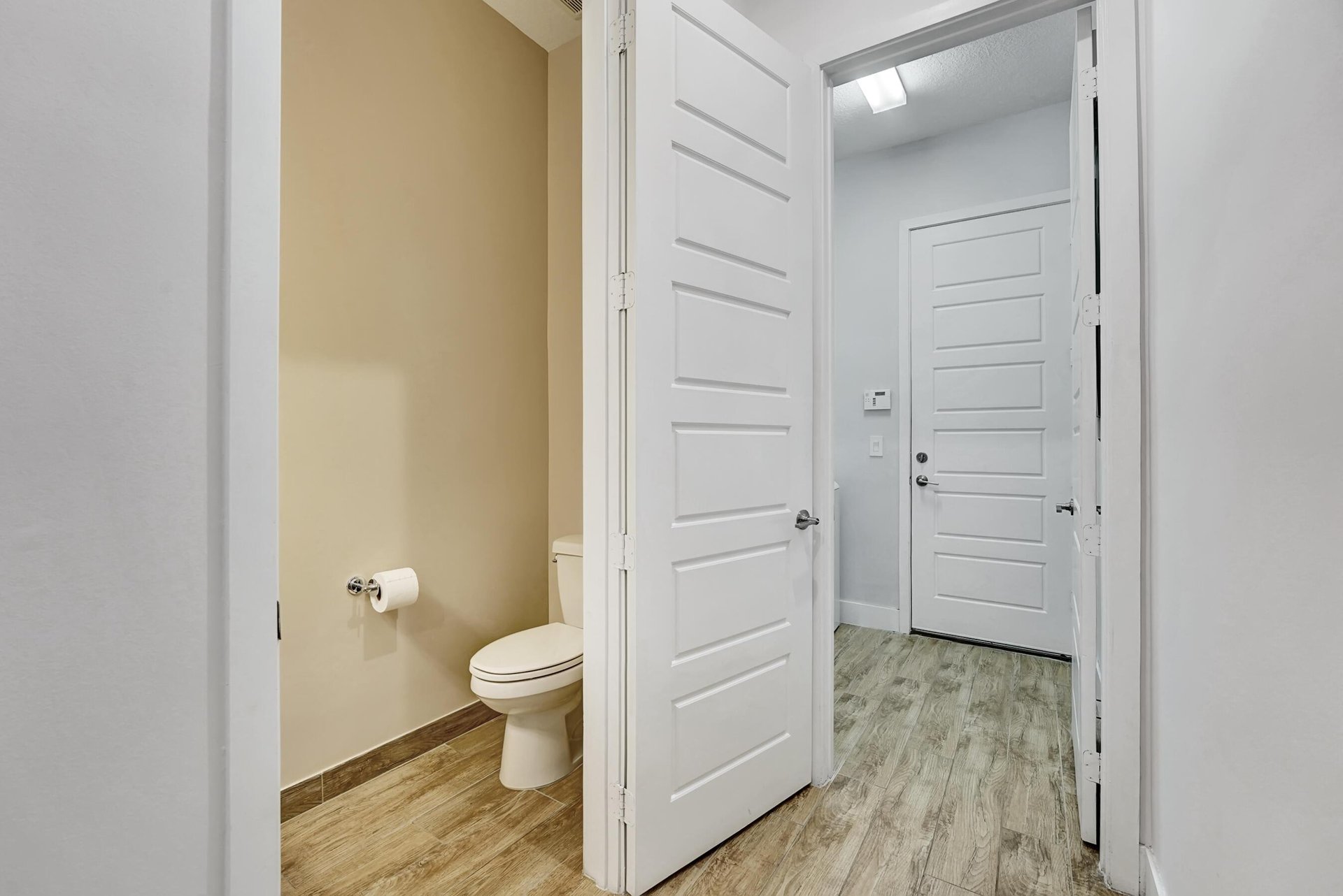Bathroom with a toilet and tan walls, viewed through an open paneled door beside a hallway.