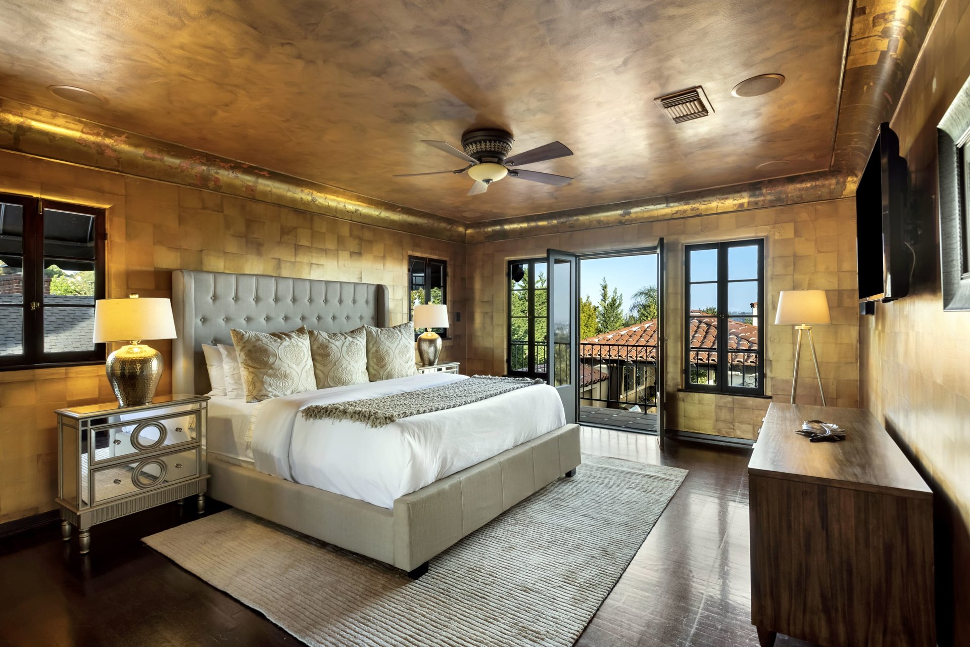 Bedroom with a tufted bed, gold-toned walls, and open doors overlooking terracotta rooftops.