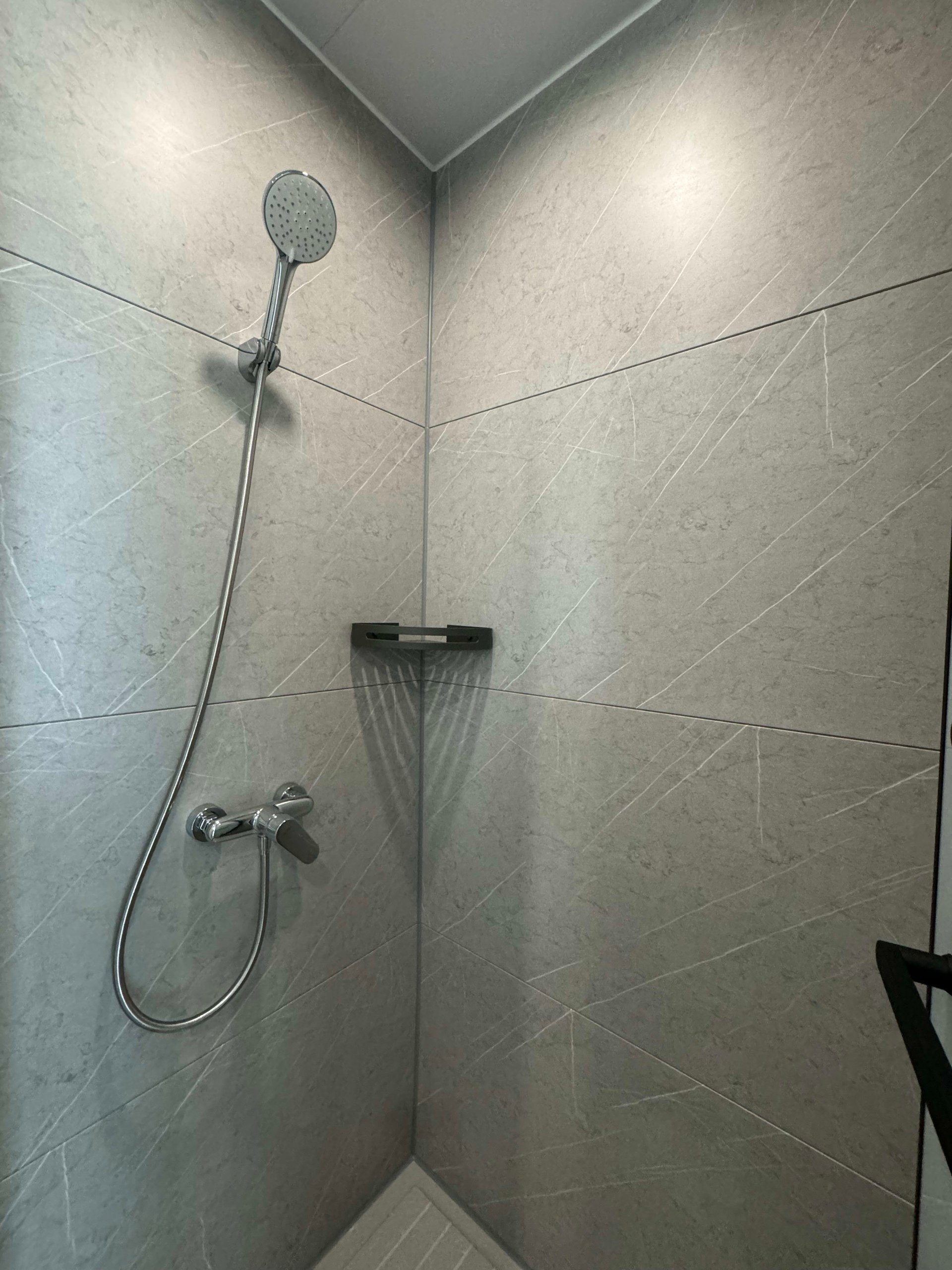 Gray tiled shower with a handheld showerhead, chrome controls, and a small black shelf.