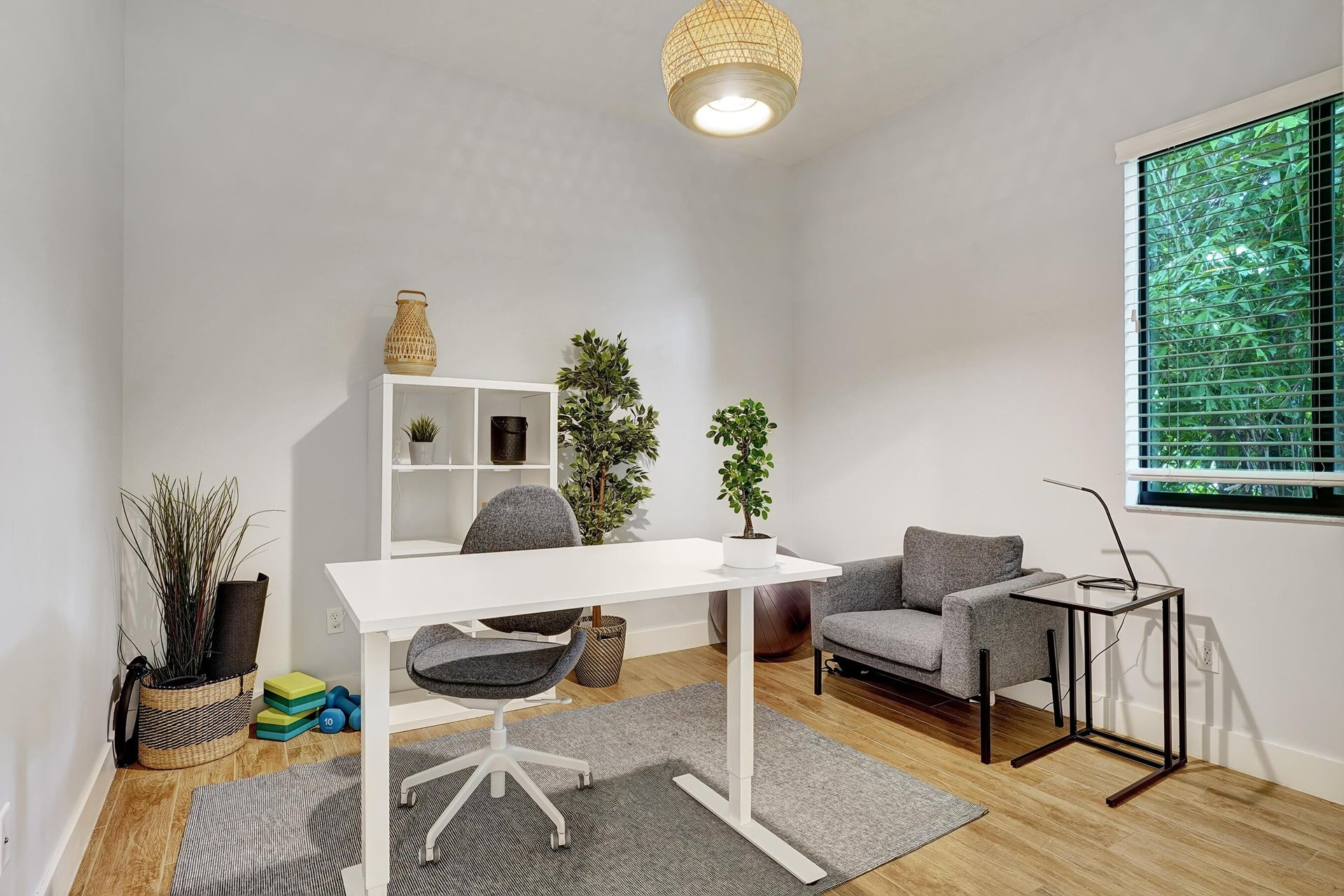 Home office with a white desk, gray armchair, indoor plants, and a large window.