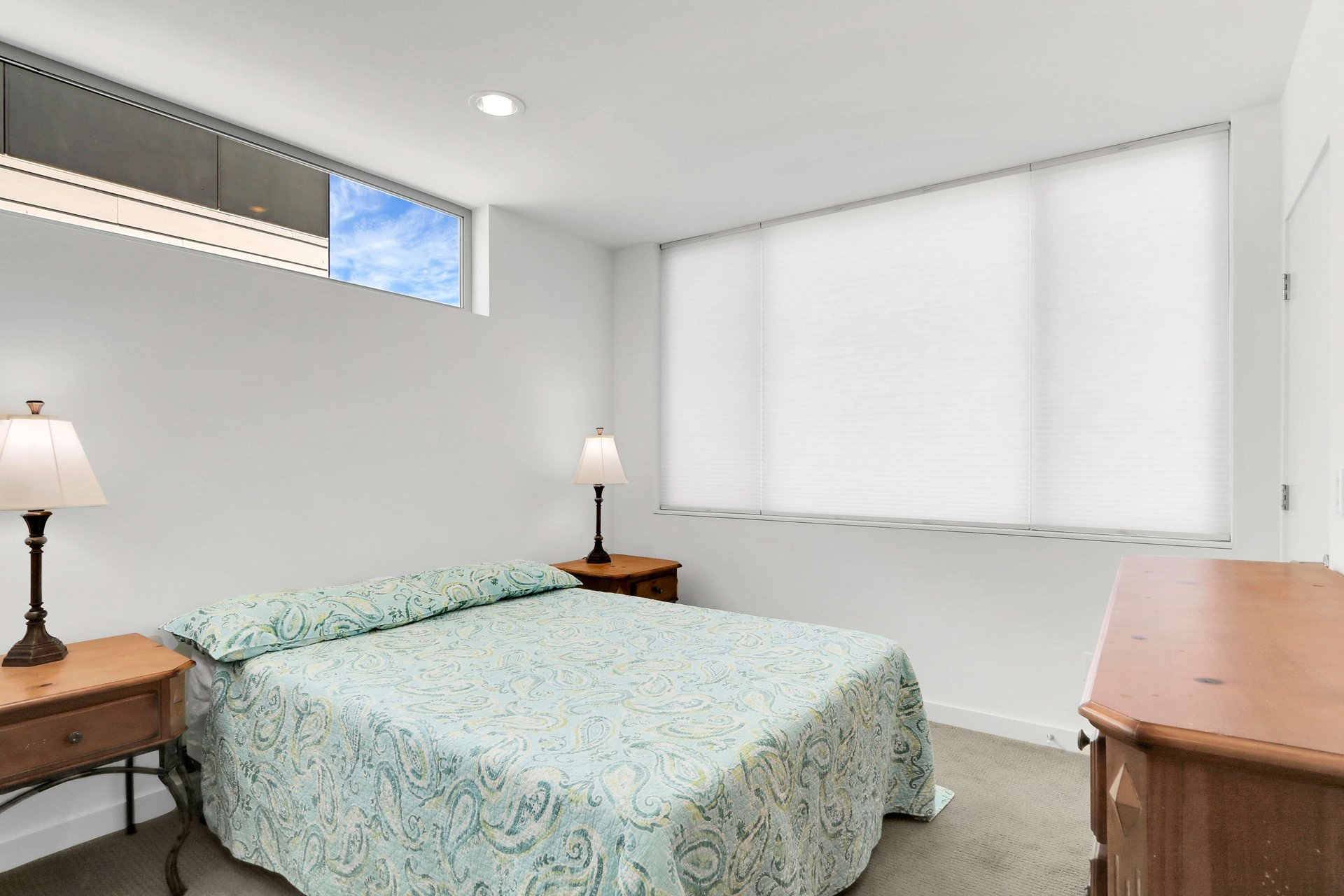 Bedroom with a patterned bedspread, wooden furniture, and high windows with closed blinds.