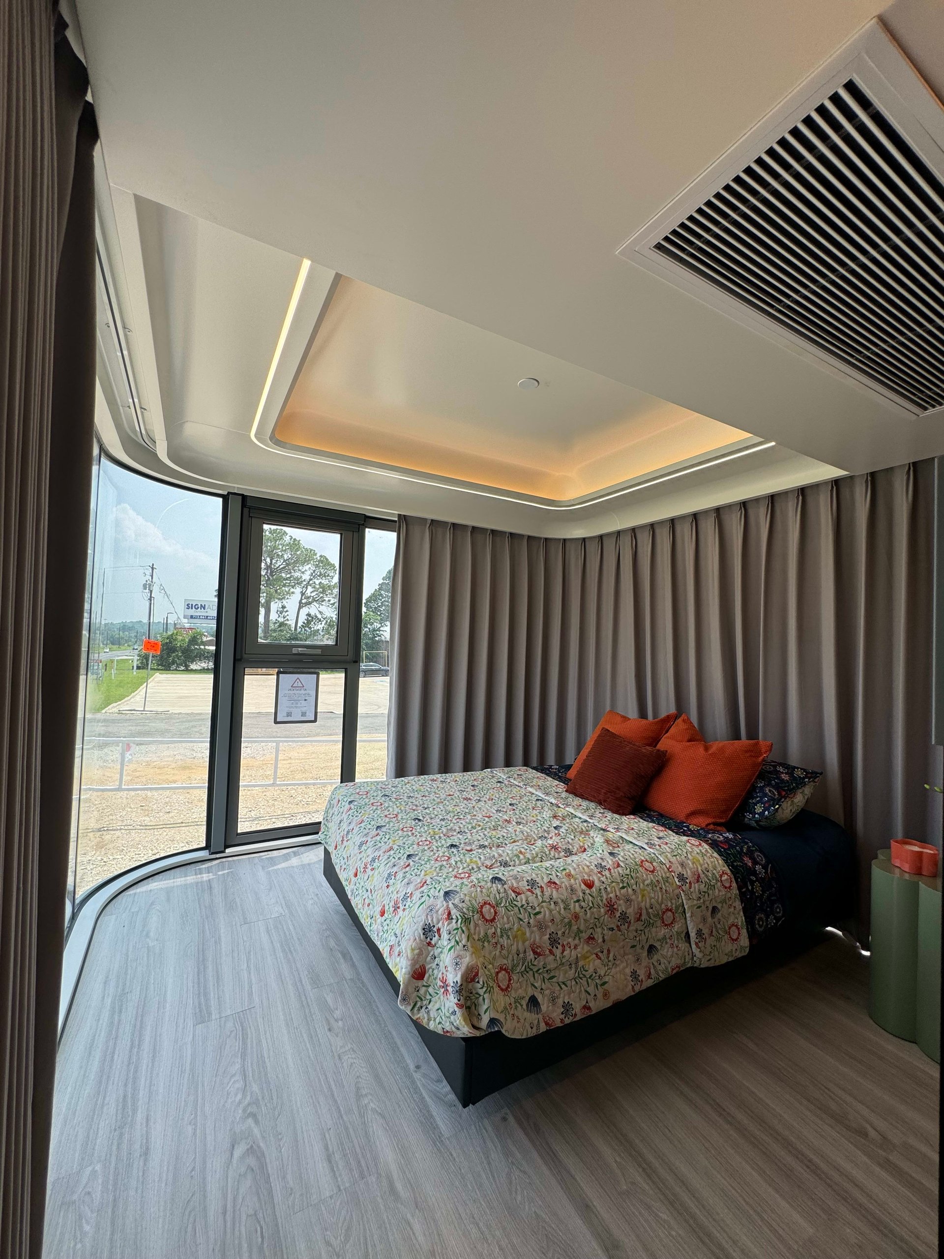 Bedroom with a floral-covered bed, orange pillows, curved windows, and warm recessed ceiling lighting.
