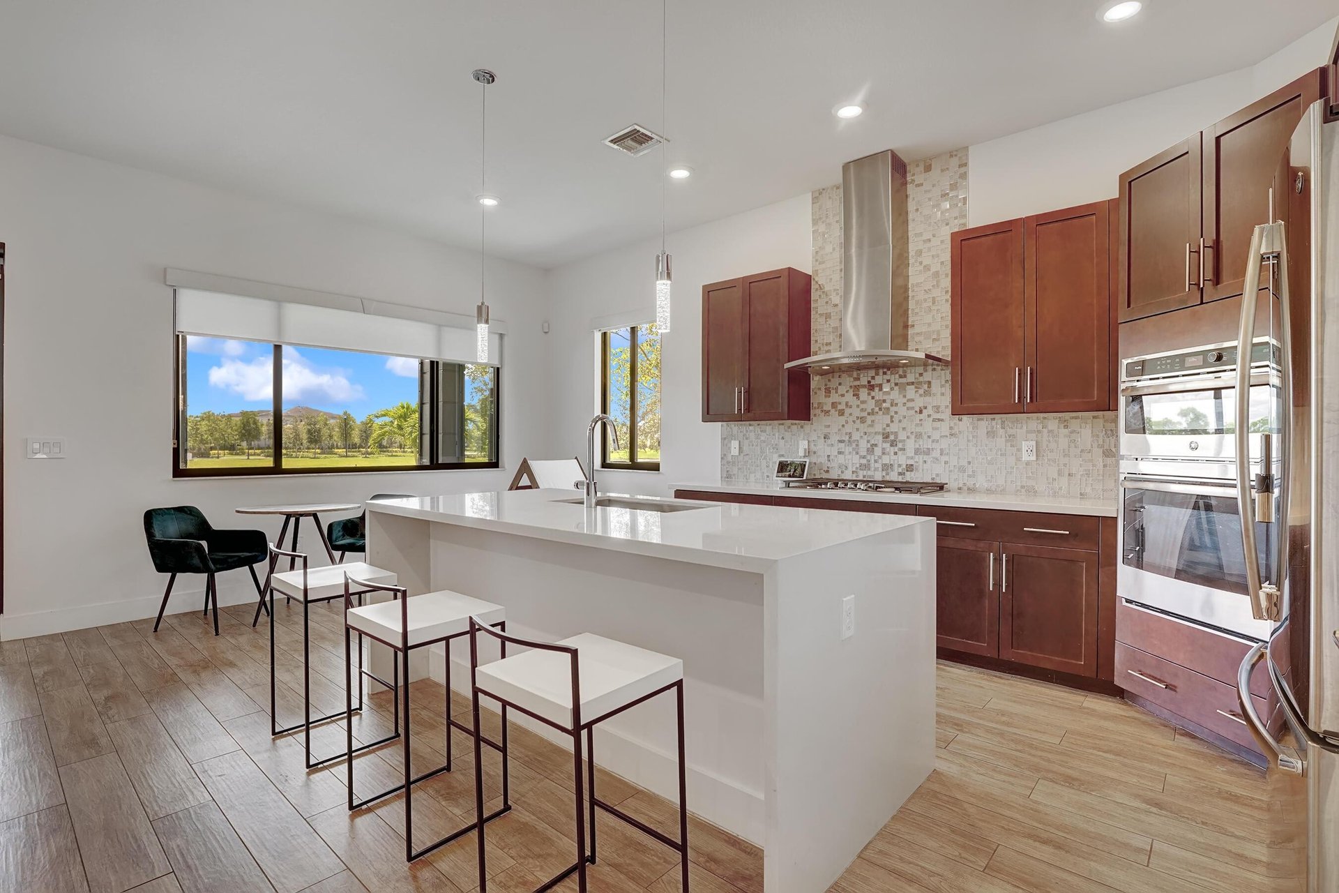 Modern kitchen with a large white island, reddish-brown cabinets, and windows overlooking a green lawn.