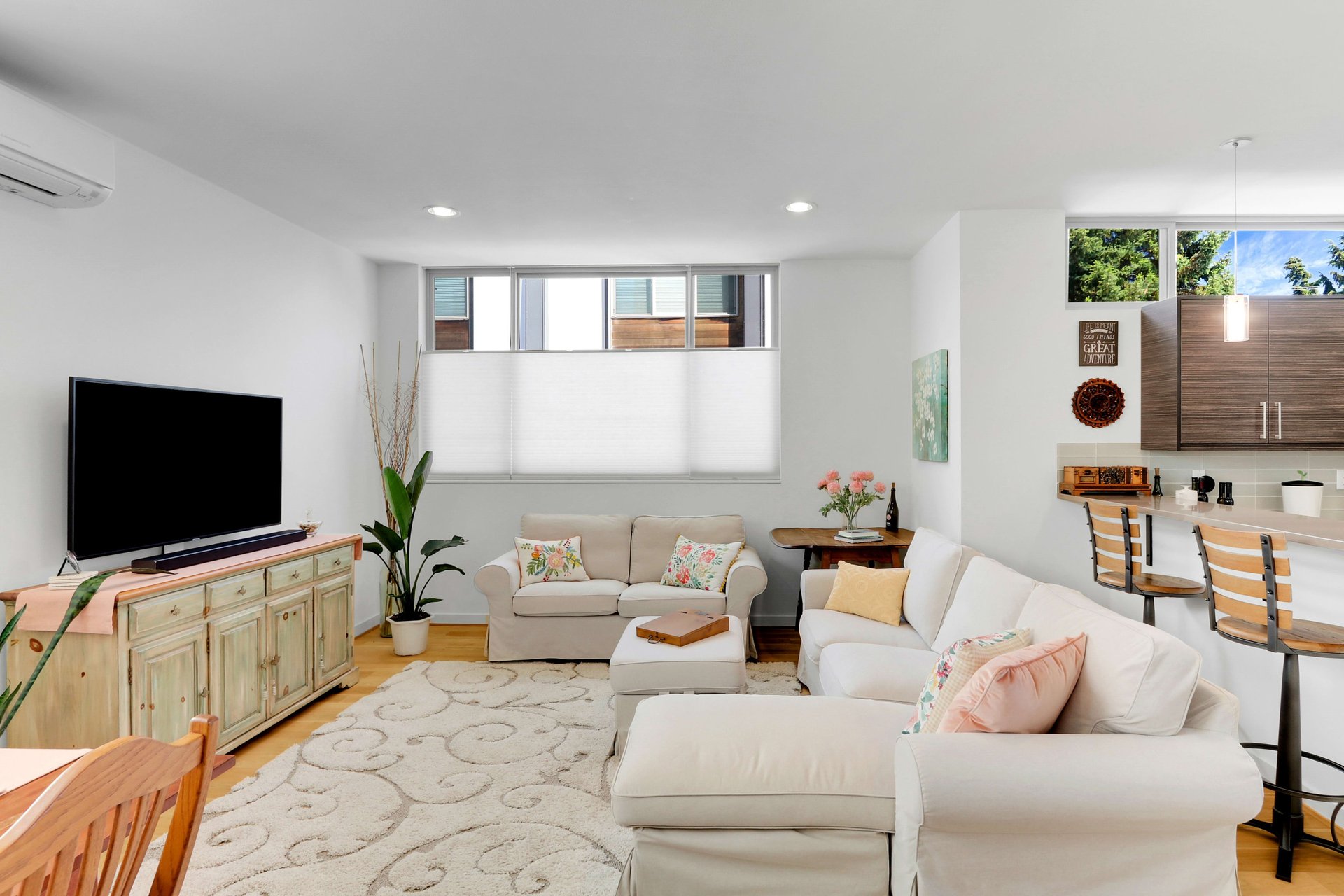 Open-plan living room with white sectional sofas, large windows, and an adjoining kitchen breakfast bar.