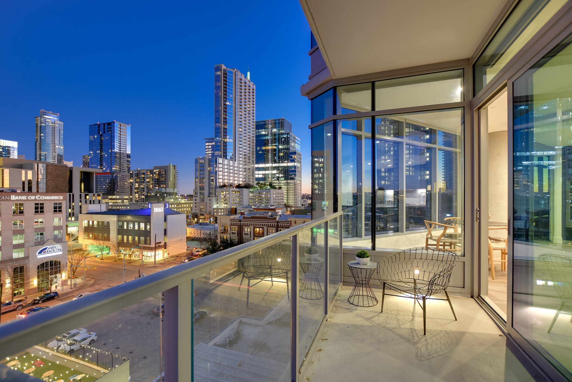 Condo balcony with glass railing and outdoor seating overlooking the downtown Austin skyline at dusk.