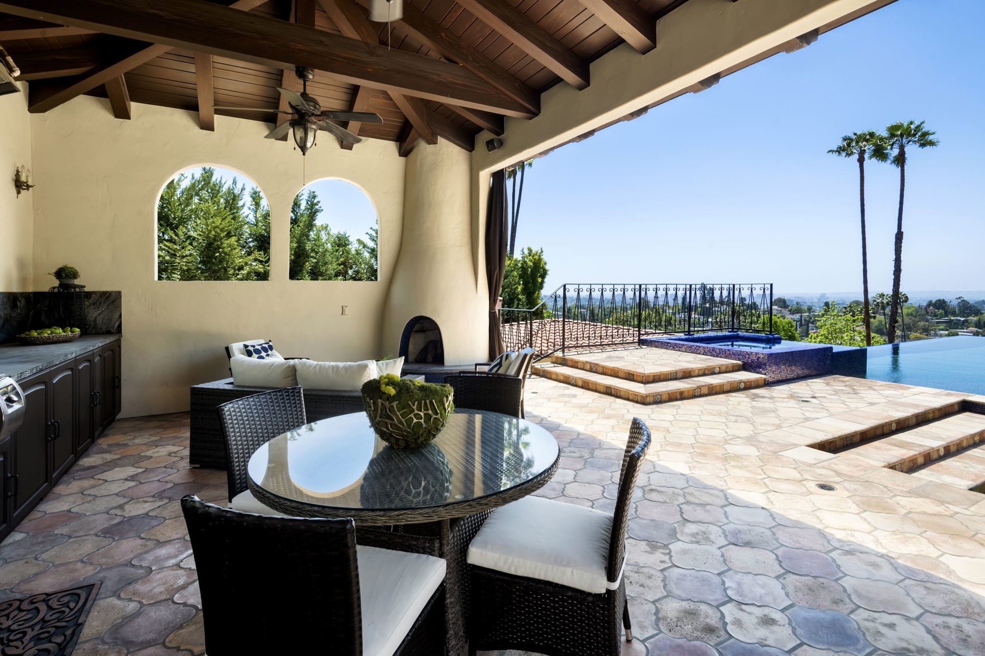 Covered patio with wicker dining set overlooking a stone terrace, infinity pool, and distant city views.